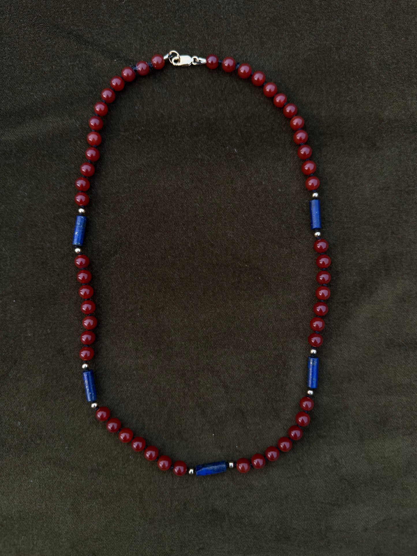 A beaded necklace with dark red round beads and alternating blue cylindrical spacer beads, displayed on a dark brown textured background.
