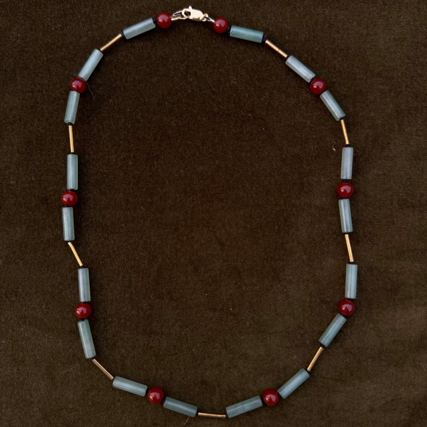 A beaded necklace featuring light blue-gray cylindrical beads, small red spherical beads, and gold-toned spacer links, arranged in an oval on a dark brown textured background.