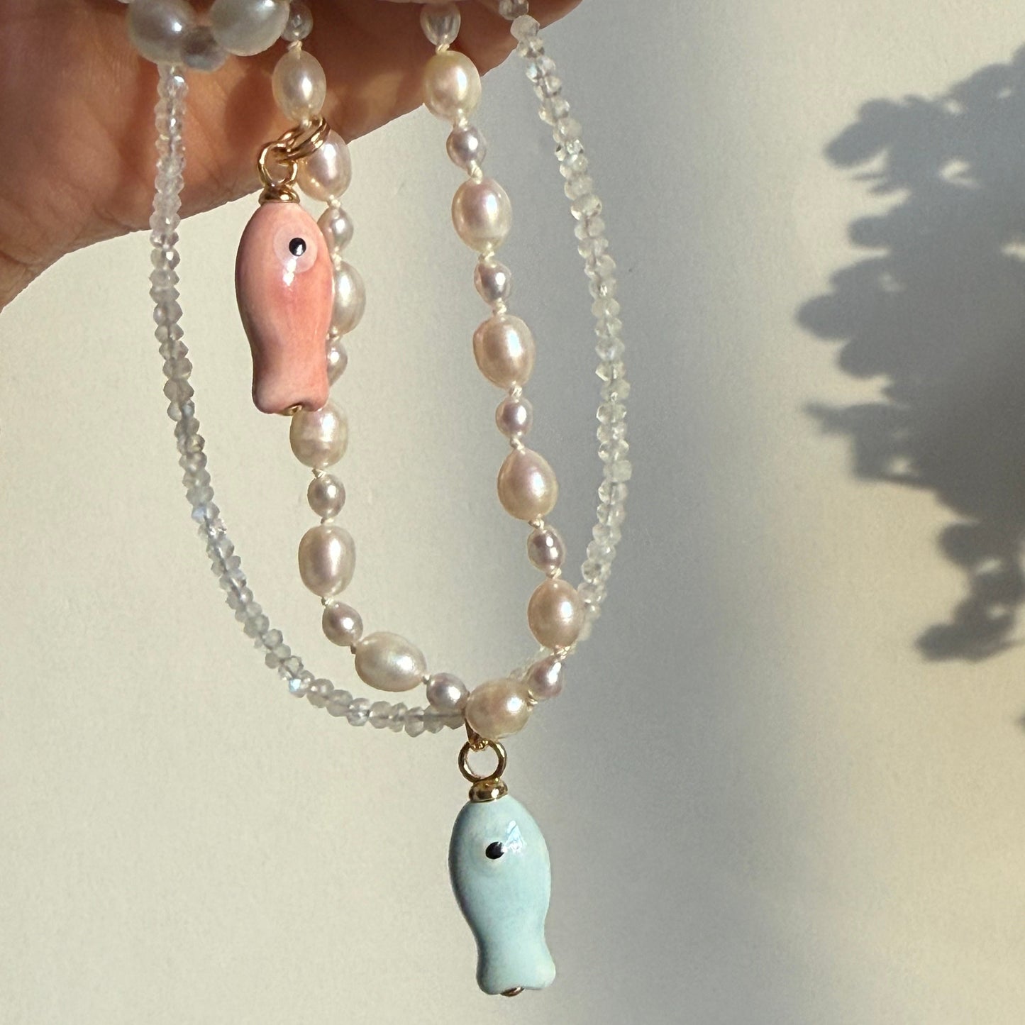 Pearl necklace with fish-shaped pendants held by a hand against a neutral background
