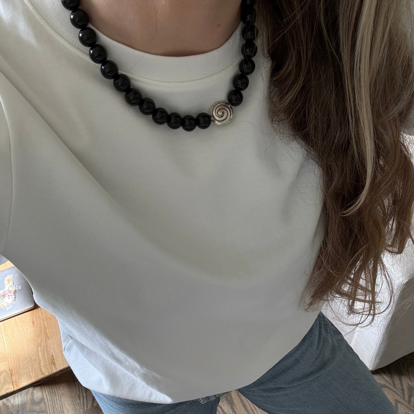 Person wearing a white shirt and black beaded necklace, sitting on a wooden floor.