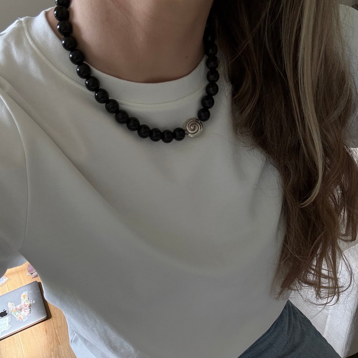 Person wearing a white tank top and black beaded necklace, sitting on a wooden floor.