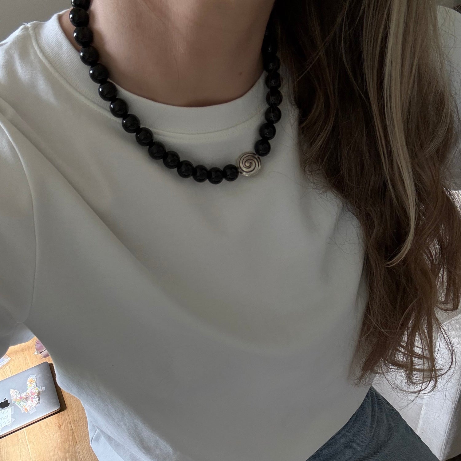 Person wearing a white tank top and black beaded necklace, sitting on a wooden floor.