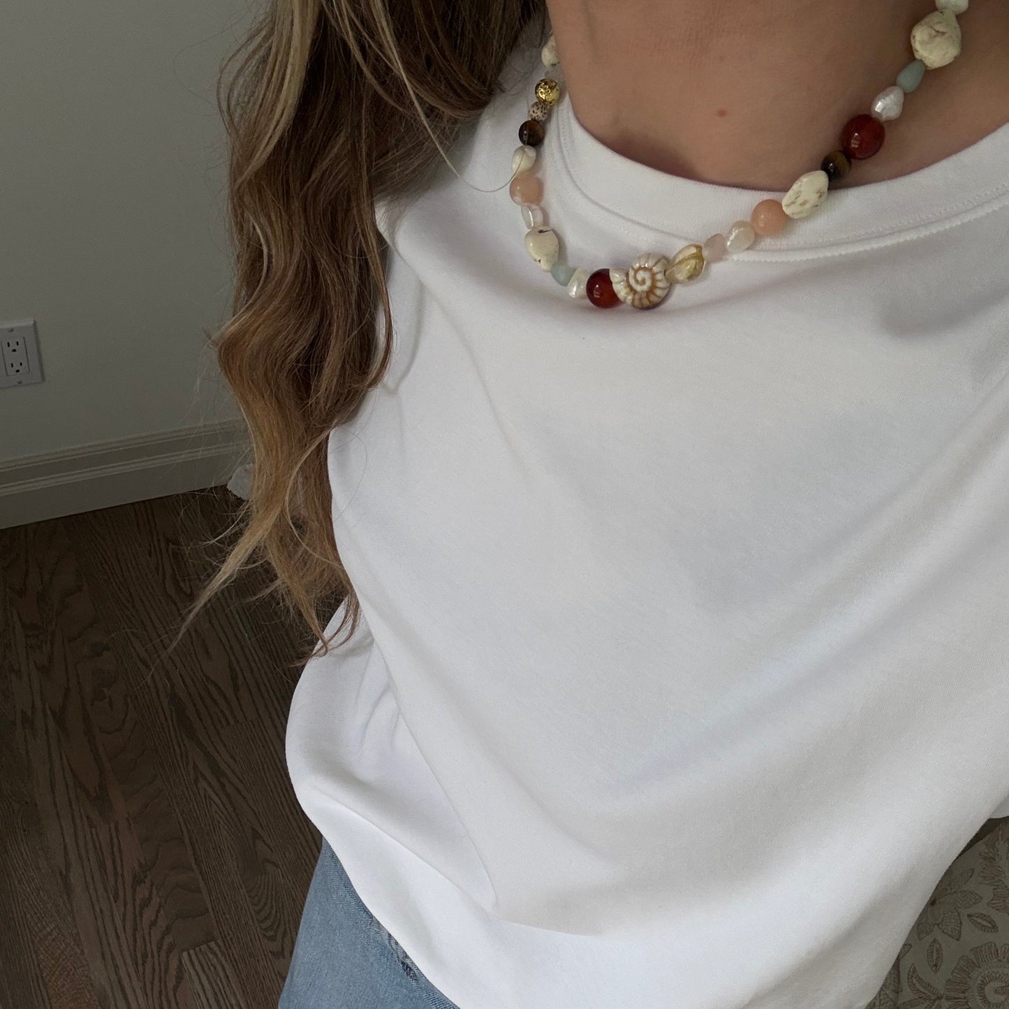 Person wearing a white shirt and blue jeans with a necklace, standing on a wooden floor.