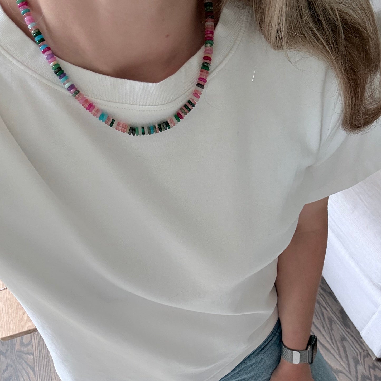 Person wearing a white sleeveless top with a colorful beaded necklace, sitting on a wooden floor.