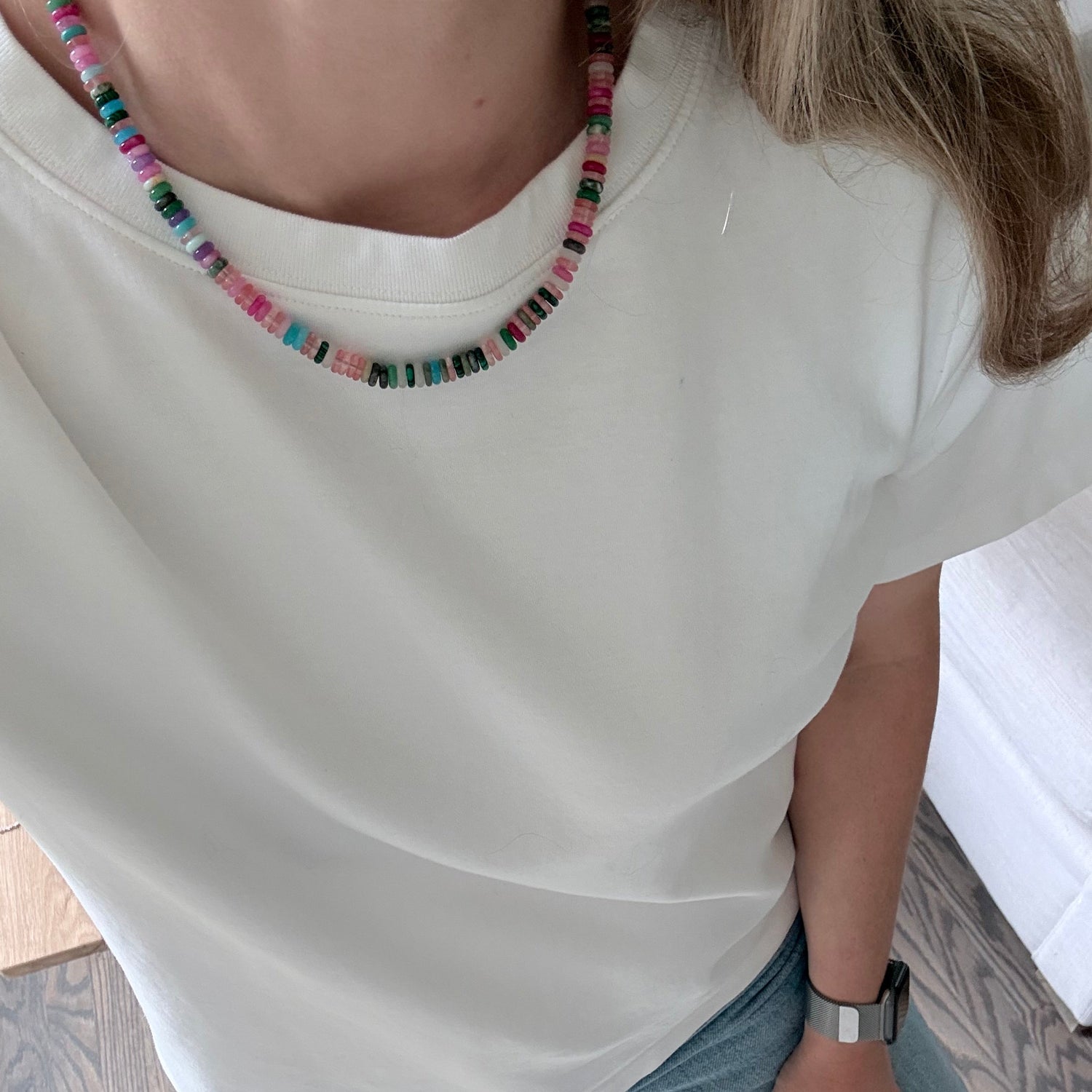 Person wearing a white sleeveless top with a colorful beaded necklace, sitting on a wooden floor.