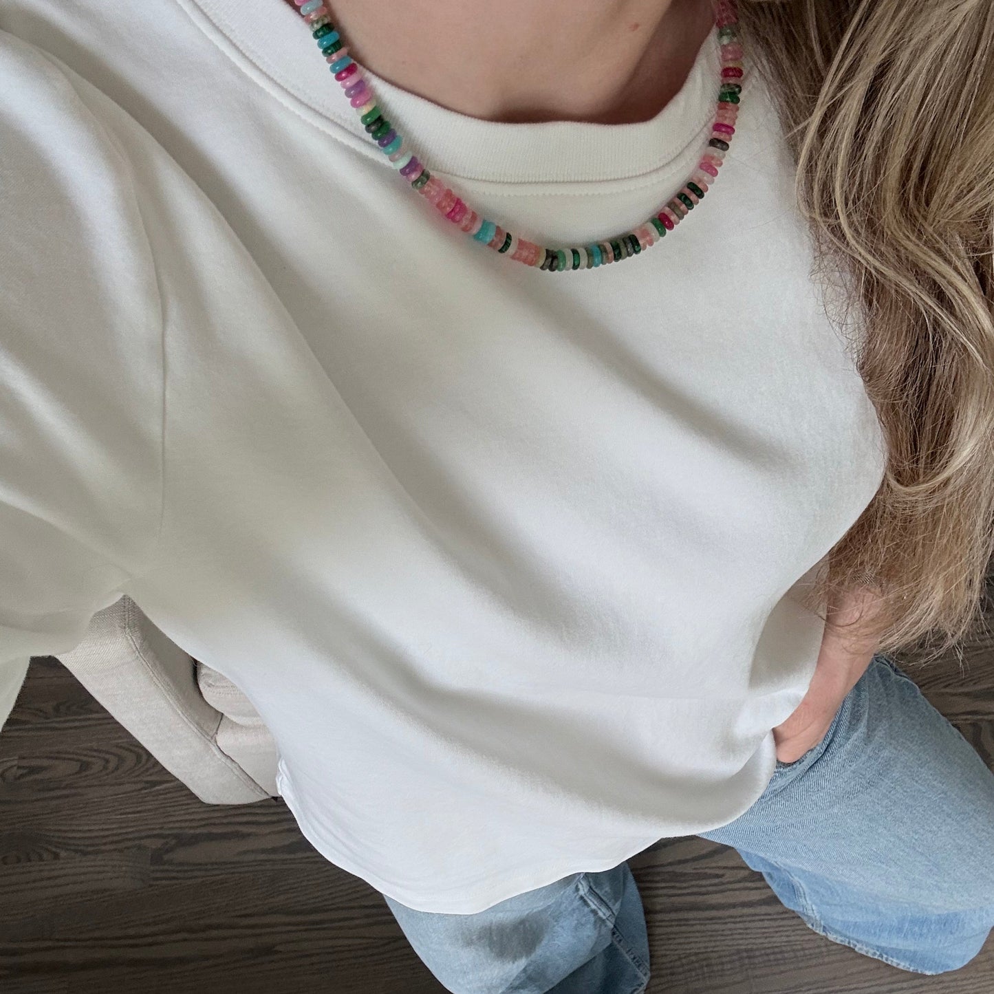 Person wearing a white hoodie and colorful beaded necklace sitting on a wooden floor.