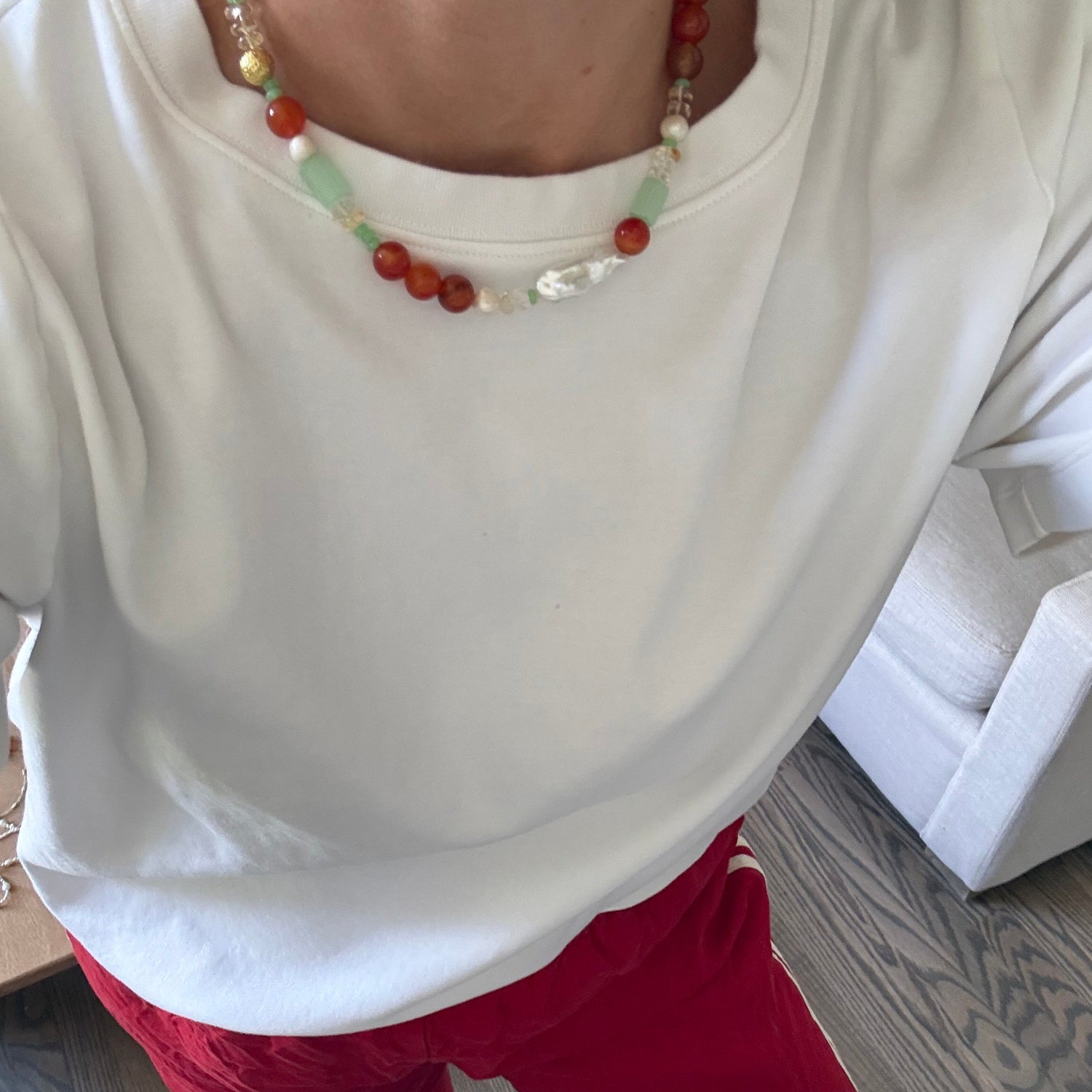 Person wearing a white shirt and red pants with a beaded necklace, sitting on a couch.