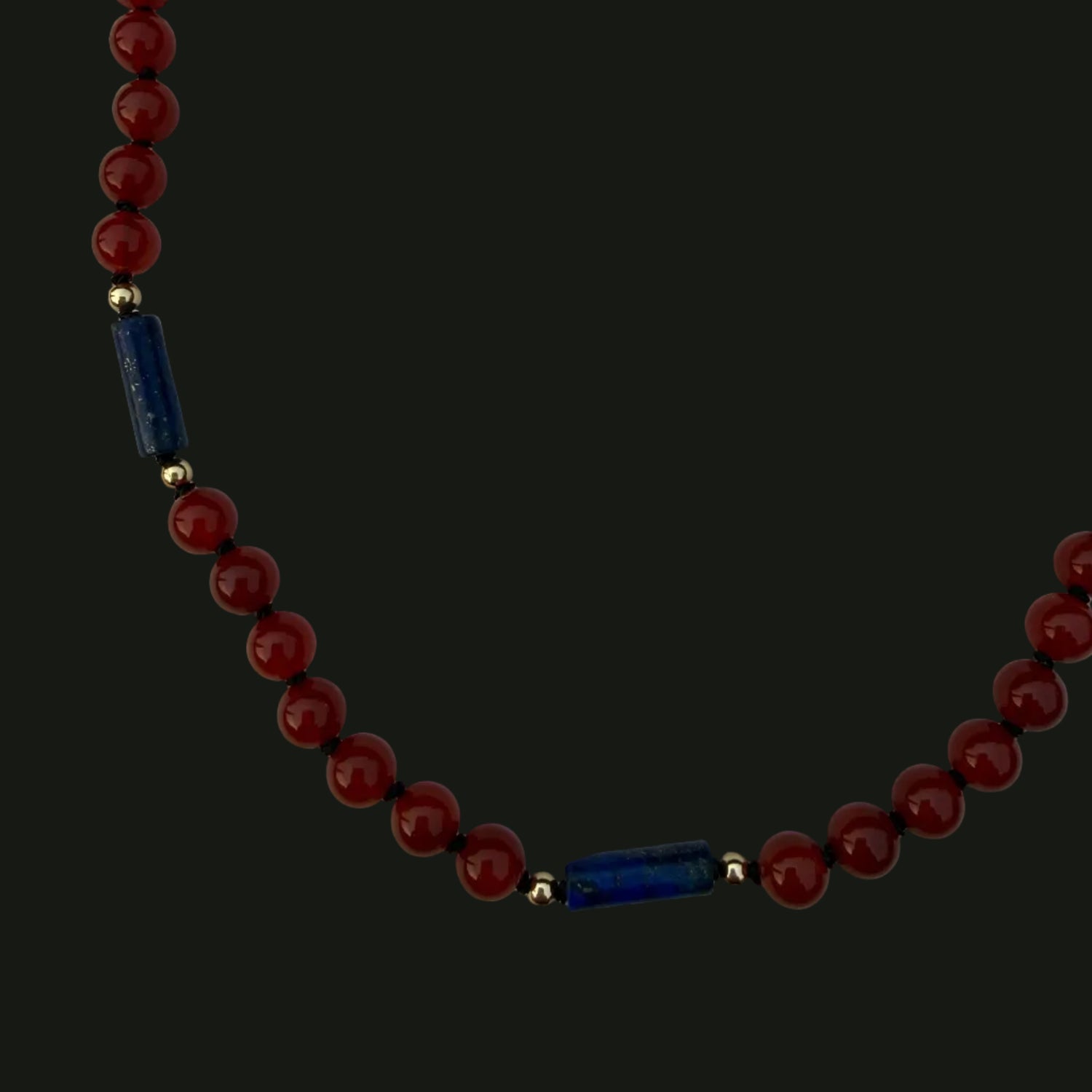 A beaded necklace featuring round dark red beads and two dark blue cylindrical spacers separated by small gold beads, displayed on a light gray background.