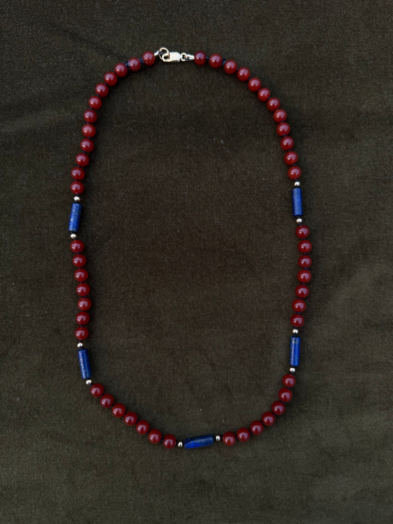 A beaded necklace with dark red round beads and alternating blue cylindrical spacer beads, displayed on a dark brown textured background.