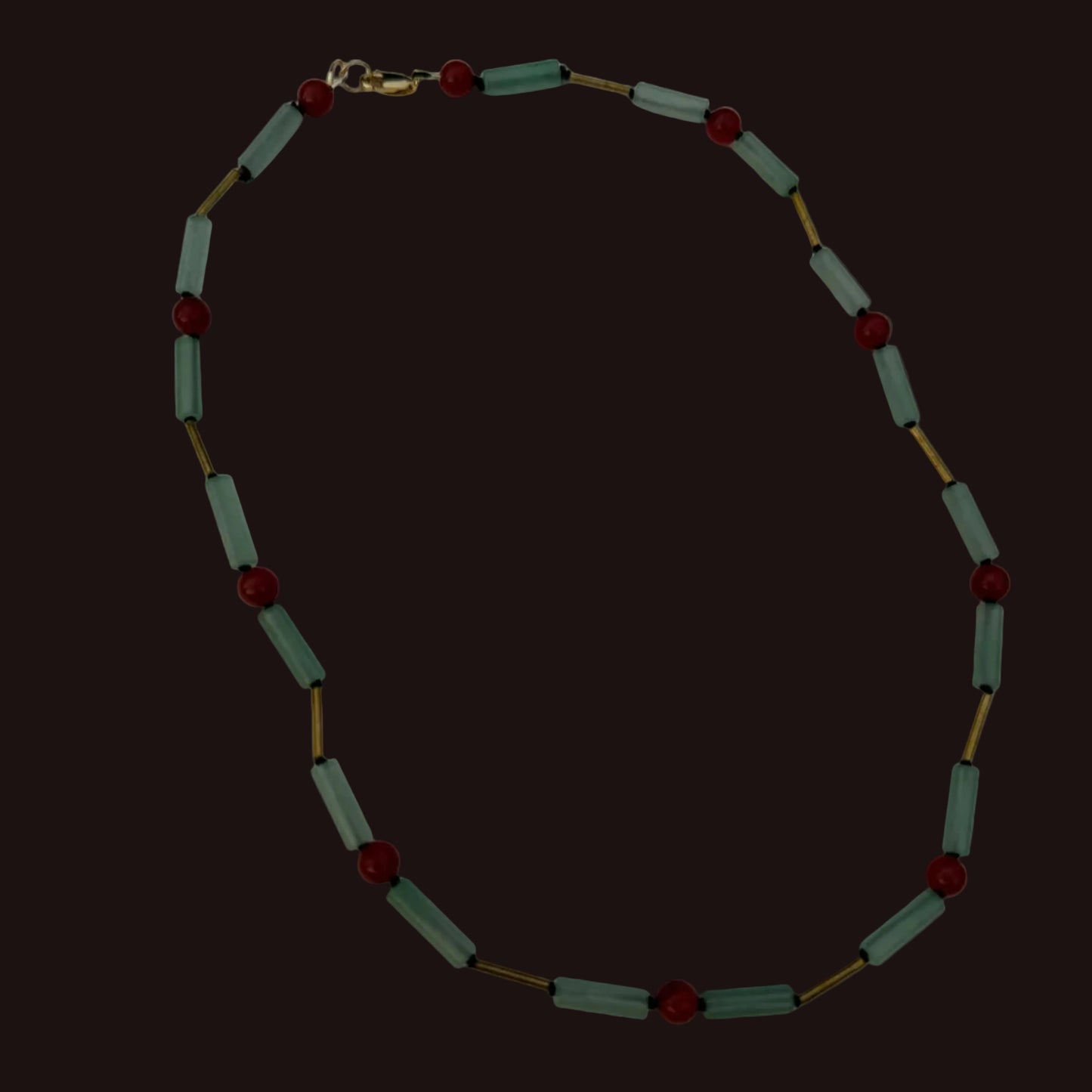 A beaded necklace featuring light sage green cylindrical beads separated by small gold-toned spacers, accented with round dark red beads, laid out on a plain white surface.