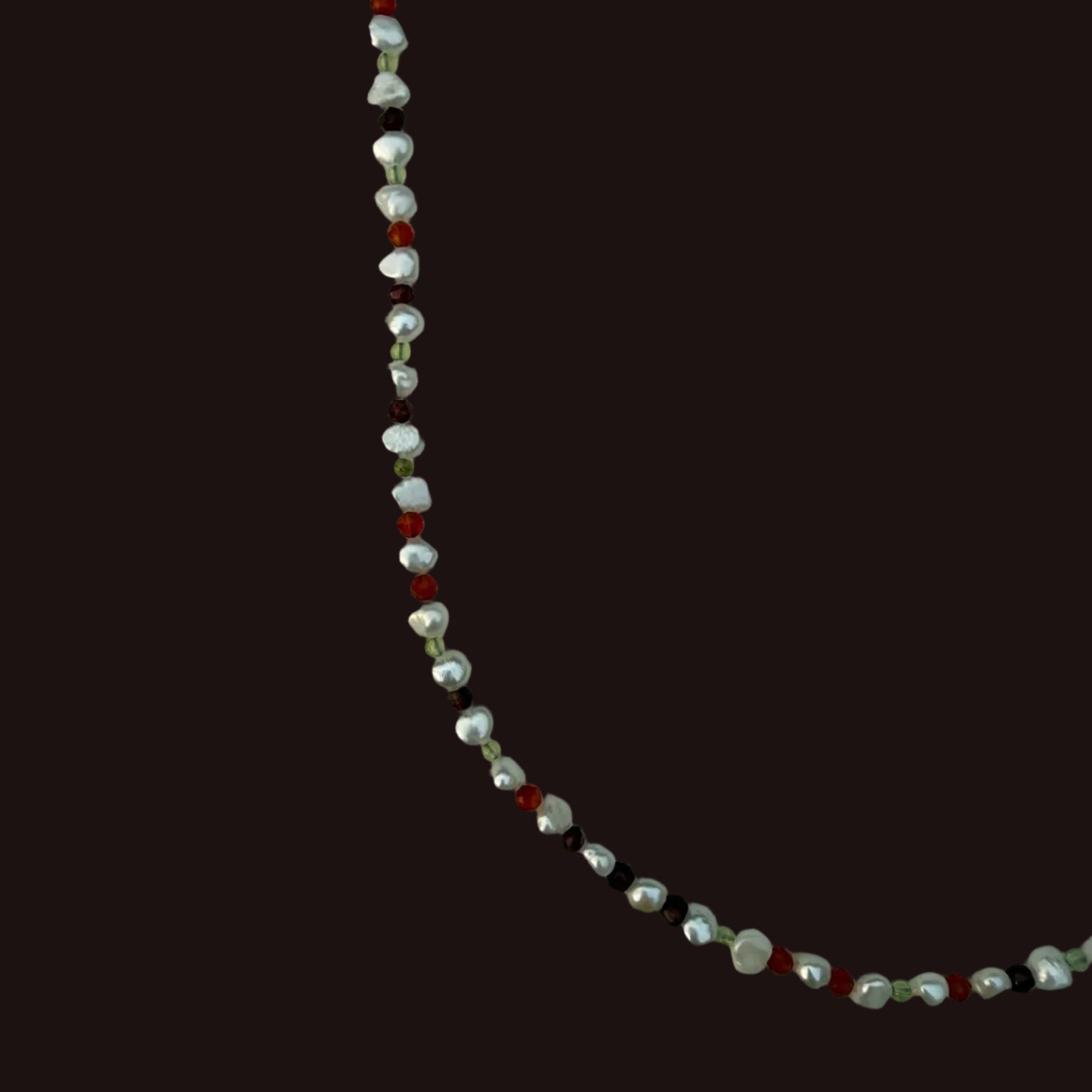 A beaded strand necklace featuring irregular white pearls, small garnet beads, and tiny peridot beads, shown against a solid dark brown background.