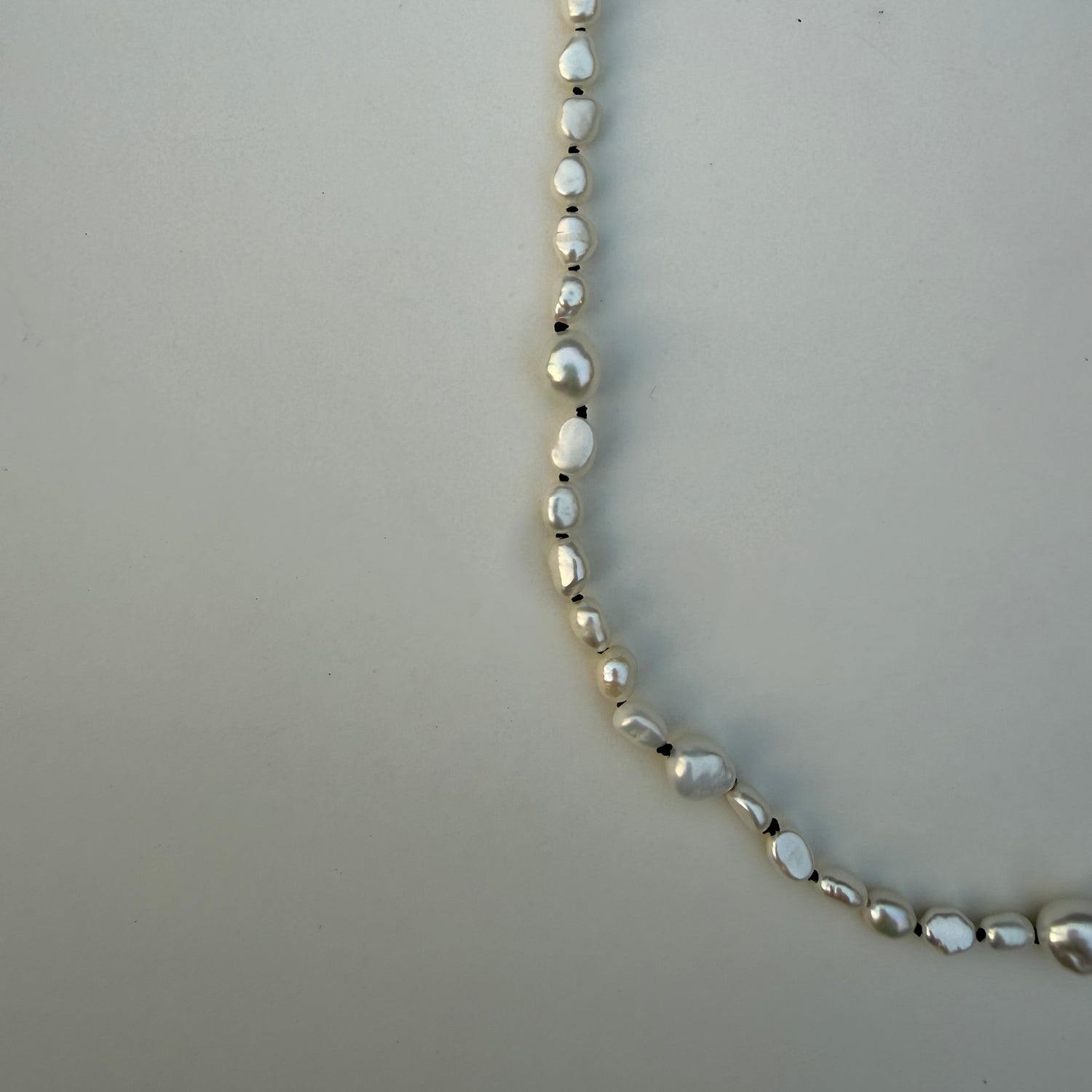 A close-up of a silver-toned beaded necklace featuring irregular, baroque-style white pearls separated by small dark spacer beads, laid out on a neutral light gray background.
