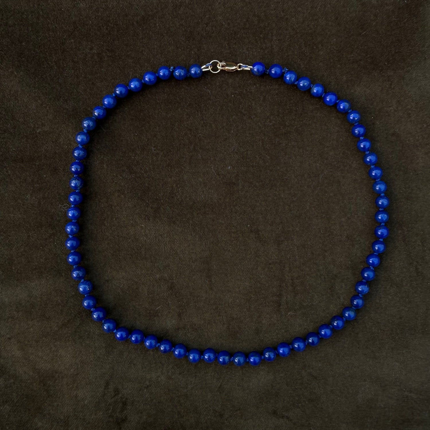 A beaded necklace made of small, glossy royal blue beads arranged in a circle, featuring a small silver lobster clasp at the top, displayed on a dark brown textured background.
