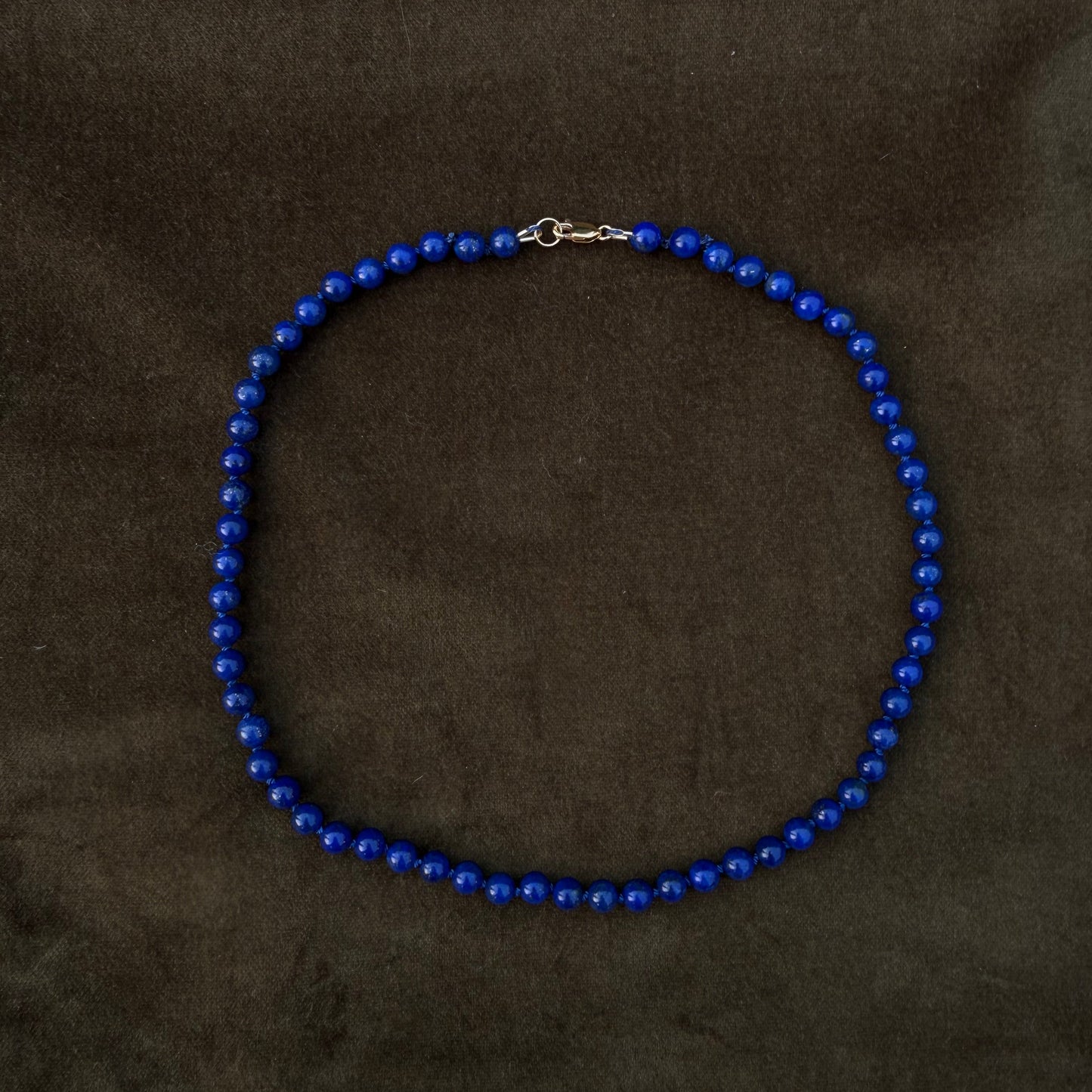 A beaded necklace made of small, glossy royal blue beads with a silver lobster clasp, displayed on a dark brown textured fabric background.