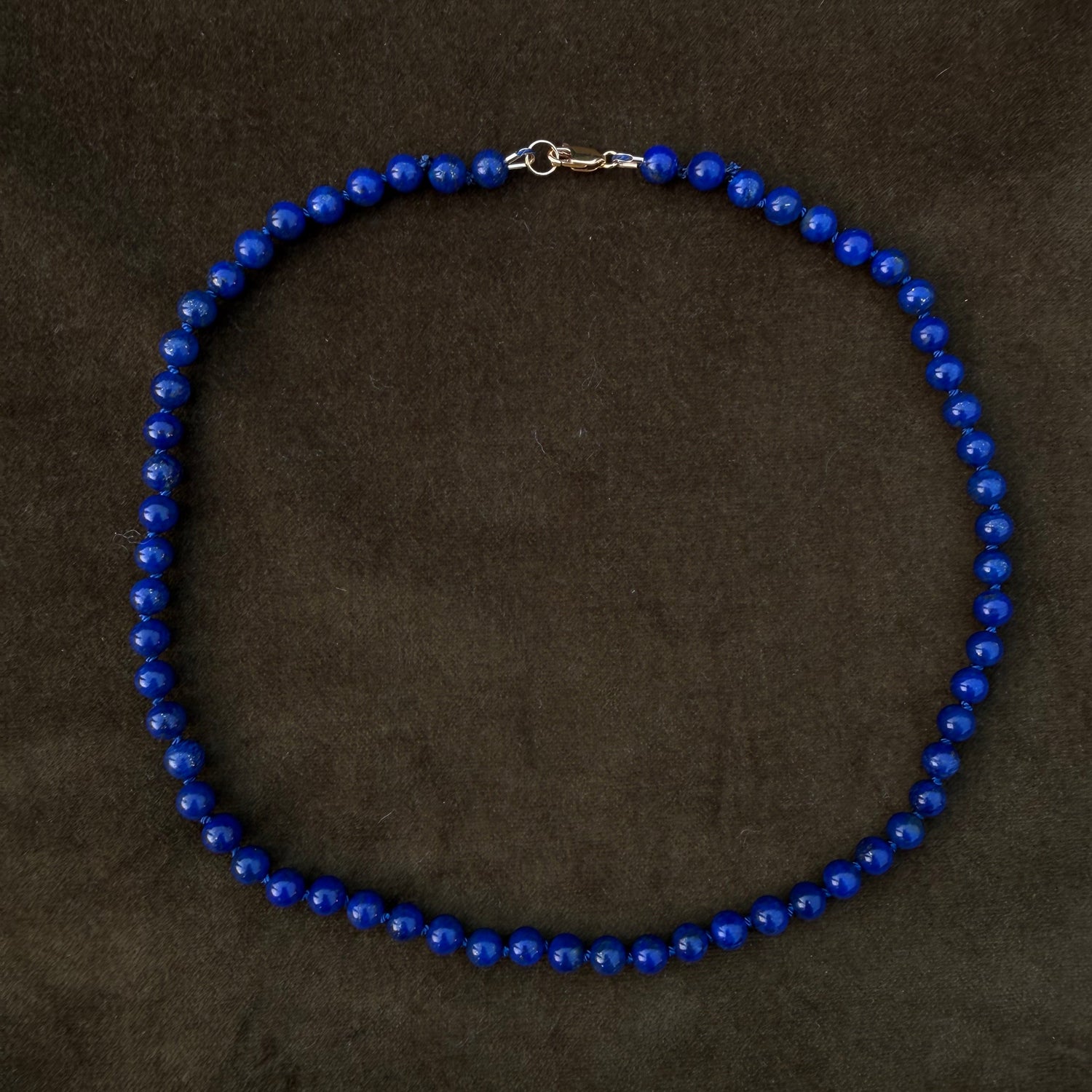 A beaded necklace made of small, glossy cobalt blue beads with a silver-tone lobster clasp, displayed on a dark brown textured background.
