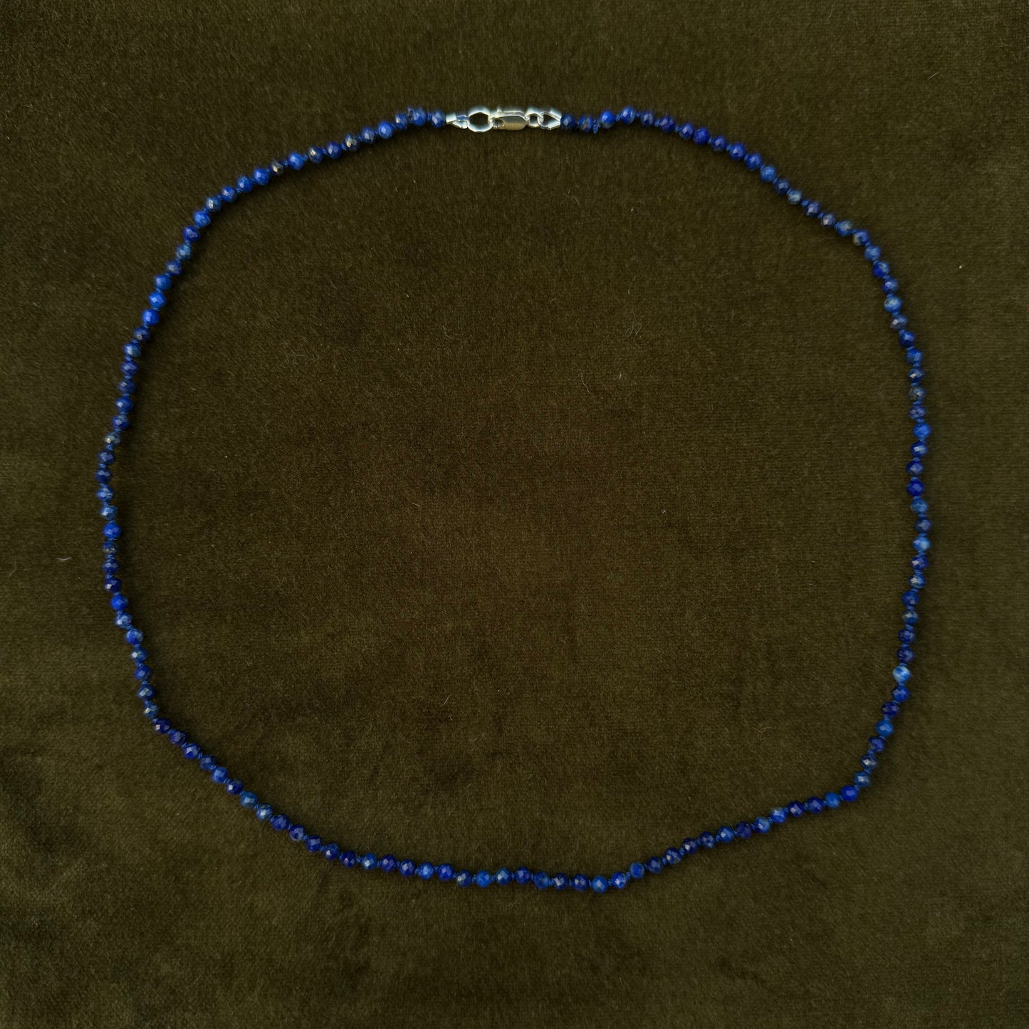 A beaded necklace made of small dark blue beads with a silver lobster clasp, displayed on a dark olive green velvet surface.