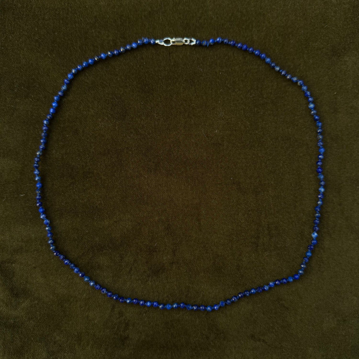 A beaded necklace made of small dark blue beads with a silver lobster clasp, displayed on a dark olive green velvet surface.
