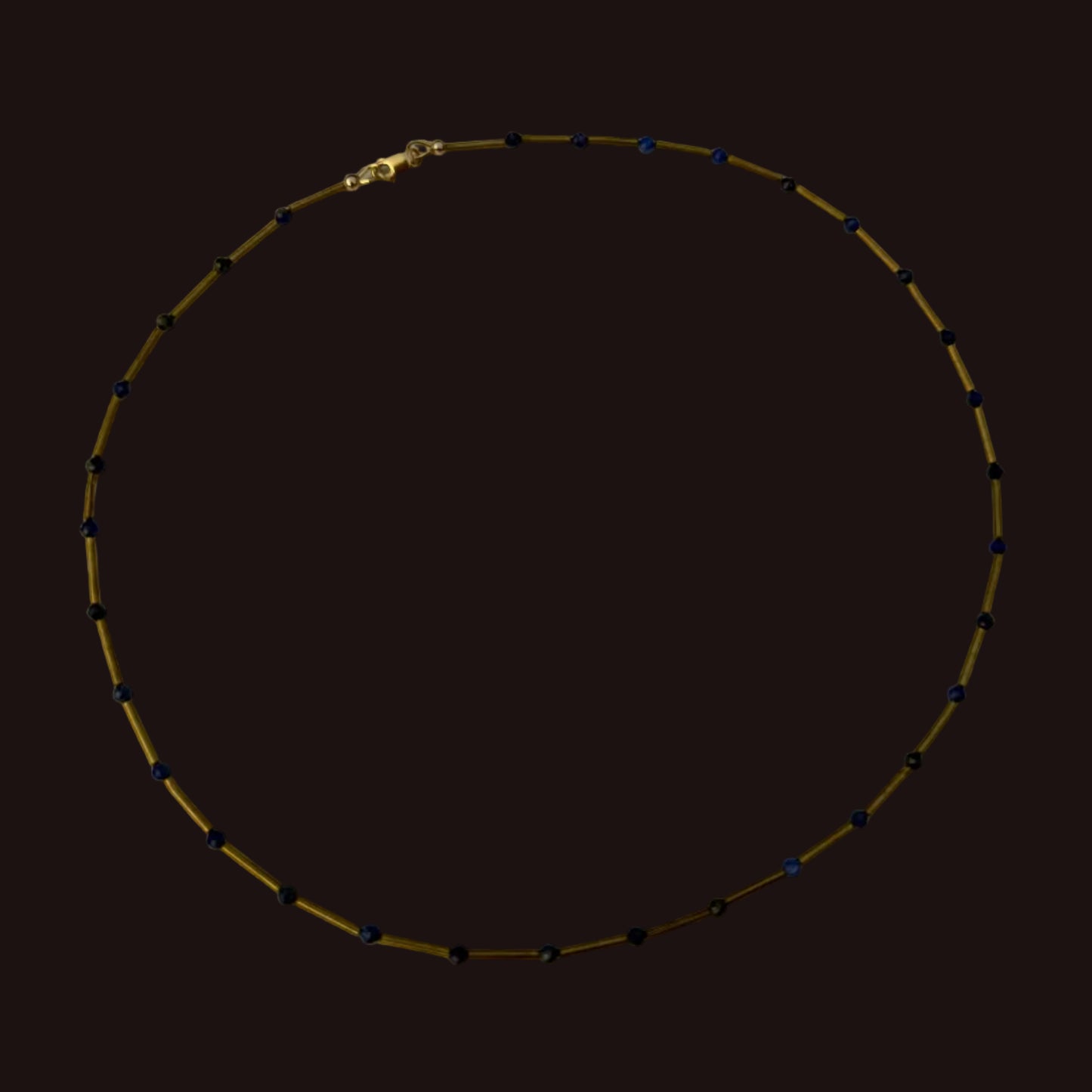 A beaded necklace with small dark blue beads spaced along a gold-toned chain, displayed in a circle on a white surface.