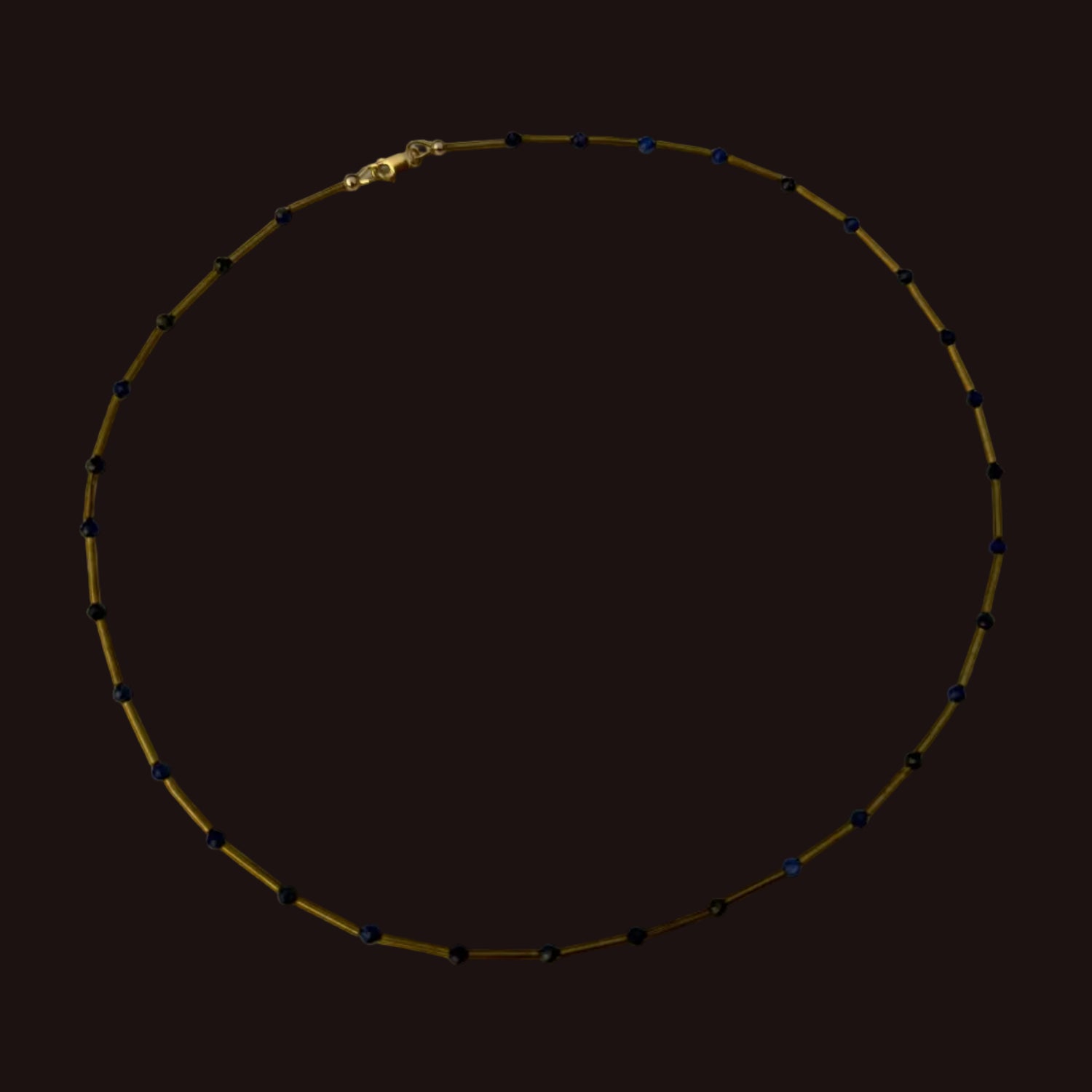 A beaded necklace with small dark blue beads spaced along a gold-toned chain, displayed in a circle on a white surface.