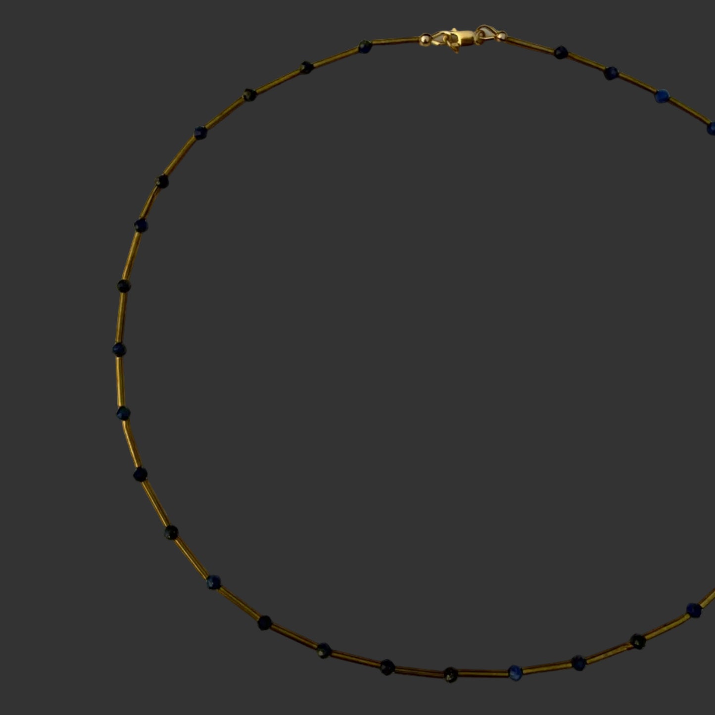 A delicate beaded necklace with small dark blue beads spaced along a thin gold-toned chain, displayed in a circle on a plain off-white surface.