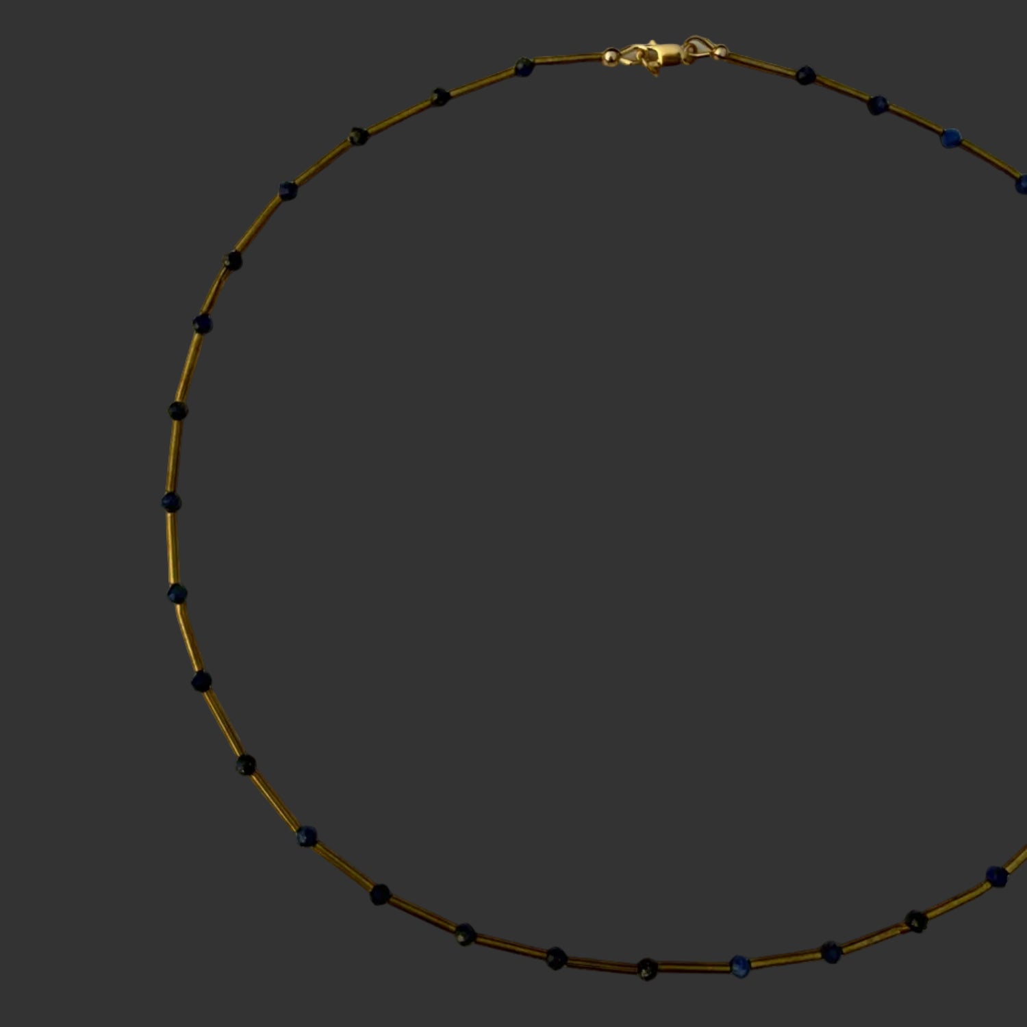 A delicate beaded necklace with small dark blue beads spaced along a thin gold-toned chain, displayed in a circle on a plain off-white surface.