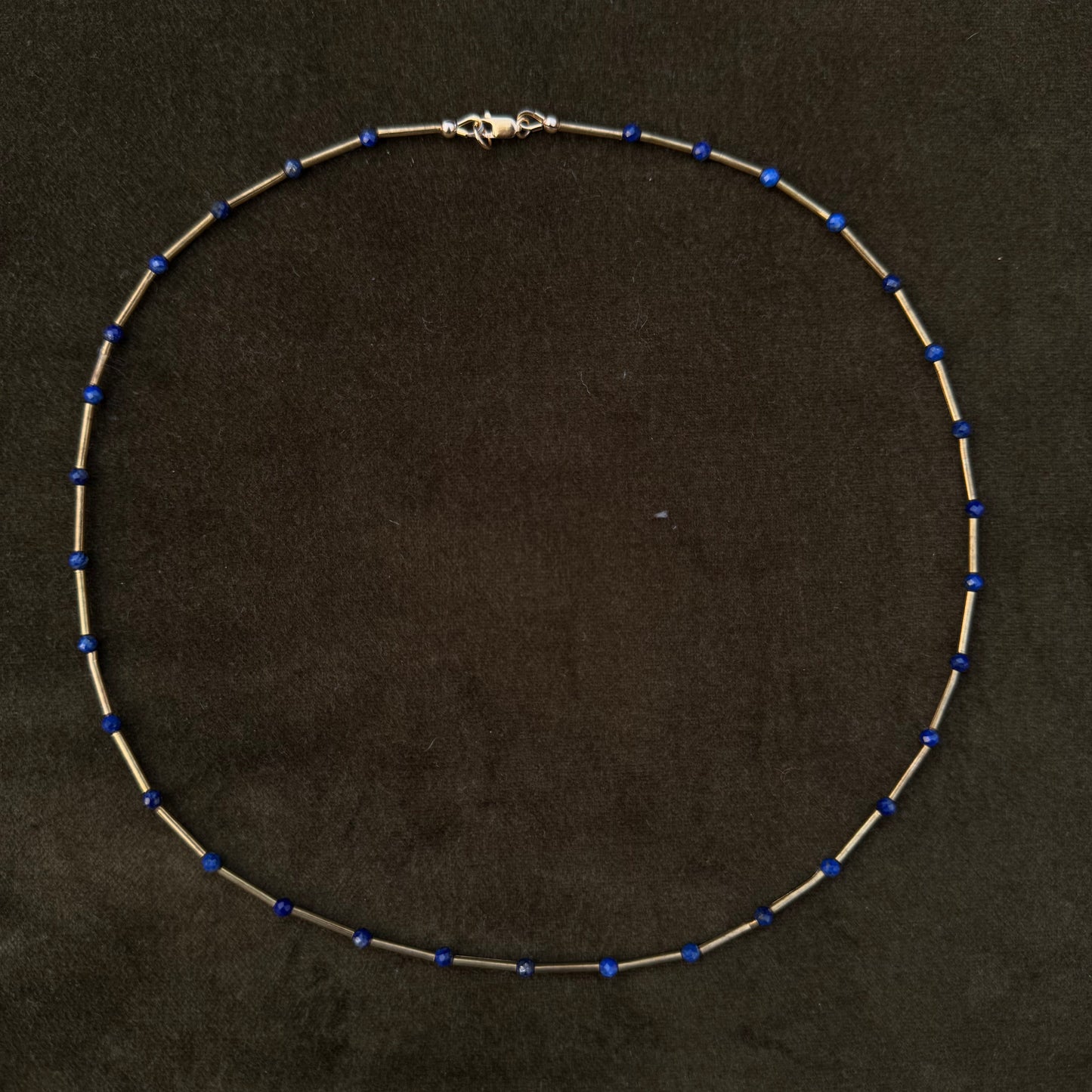 A delicate beaded necklace with small blue beads spaced along a thin silver-toned chain, displayed on a dark textured background.