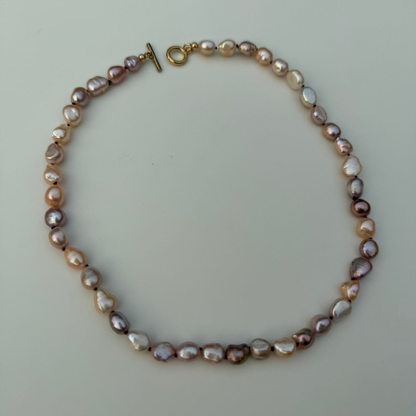 A baroque pearl necklace with irregular, polished beads in shades of lavender, pink, and cream, featuring a gold toggle clasp, displayed on a light gray background.