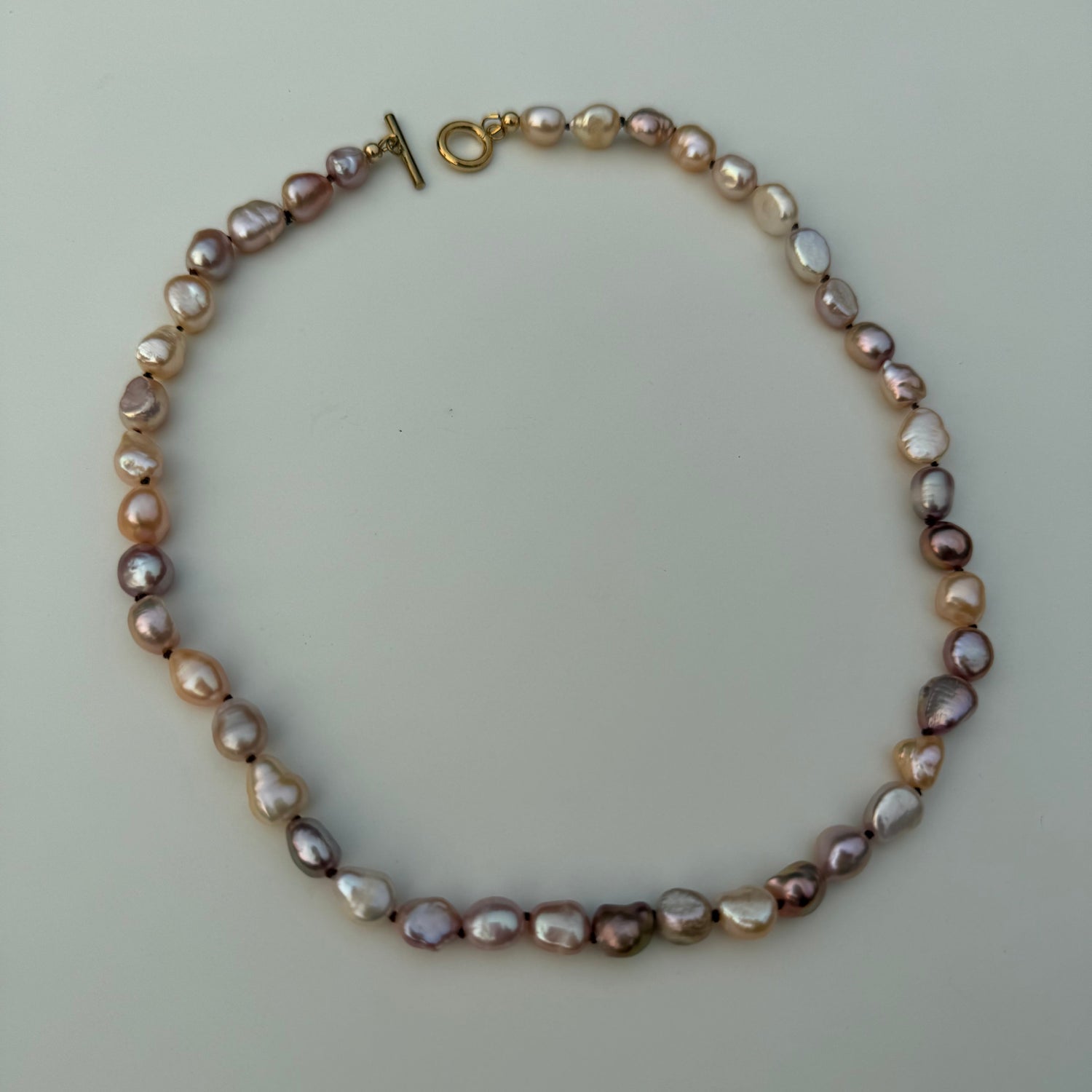 A baroque pearl necklace with irregular, polished beads in shades of lavender, pink, and cream, featuring a gold toggle clasp, displayed on a light gray background.