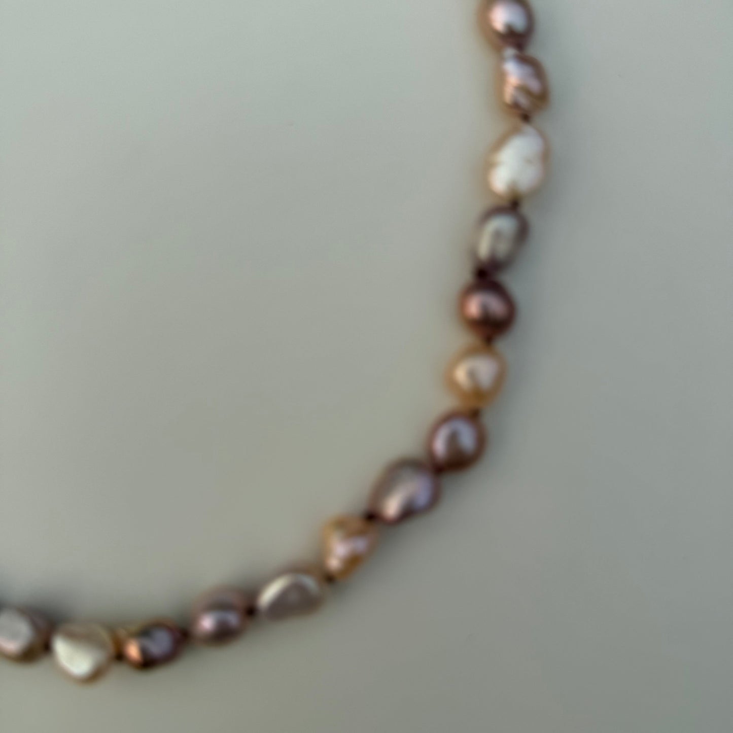 A close-up of a baroque pearl necklace with irregular, polished beads in shades of iridescent lavender, bronze, and cream, arranged in a curved line against a solid light gray background.