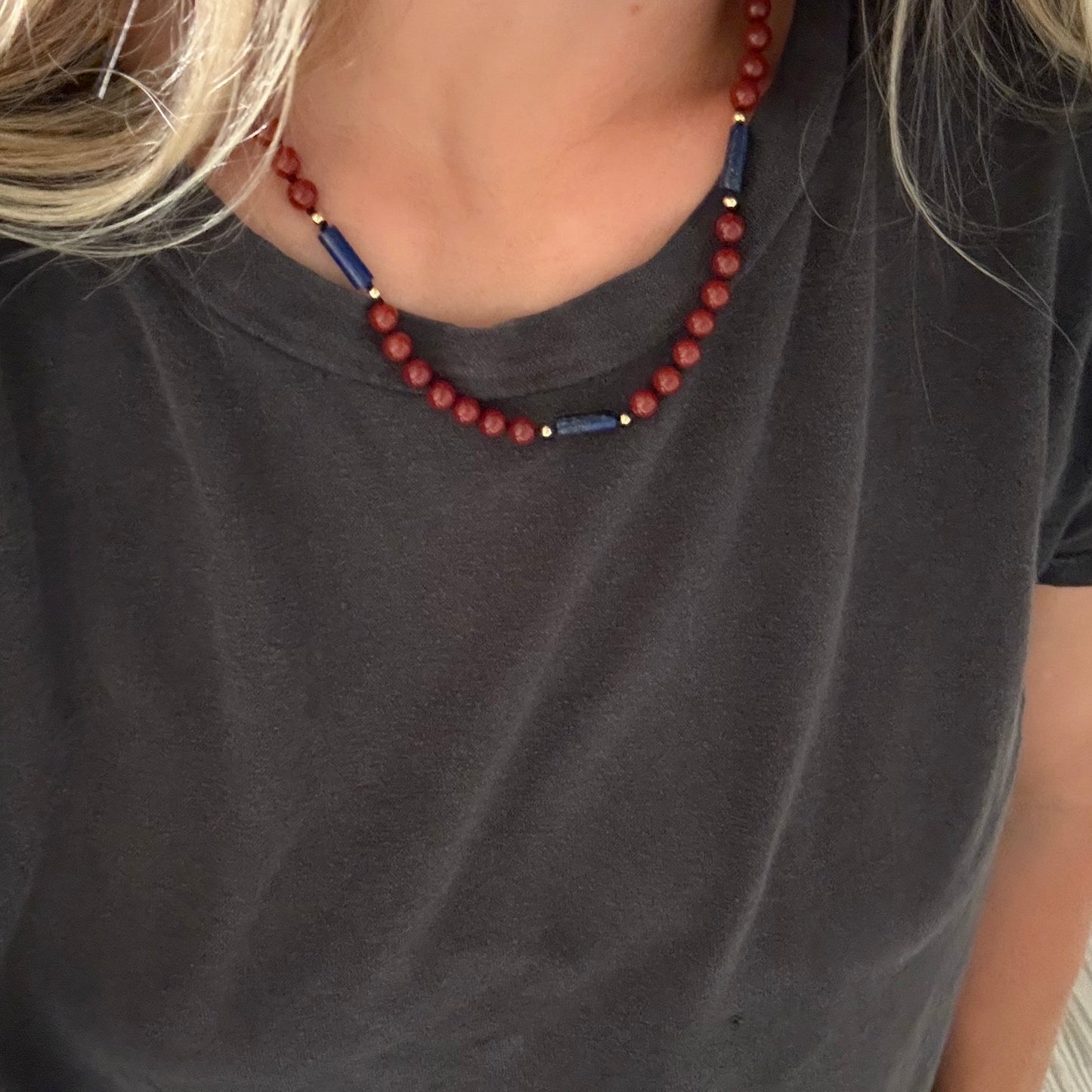 A close-up of a person wearing a beaded necklace with red round beads, blue cylindrical beads, and small gold spacer beads, paired with a dark gray t-shirt.