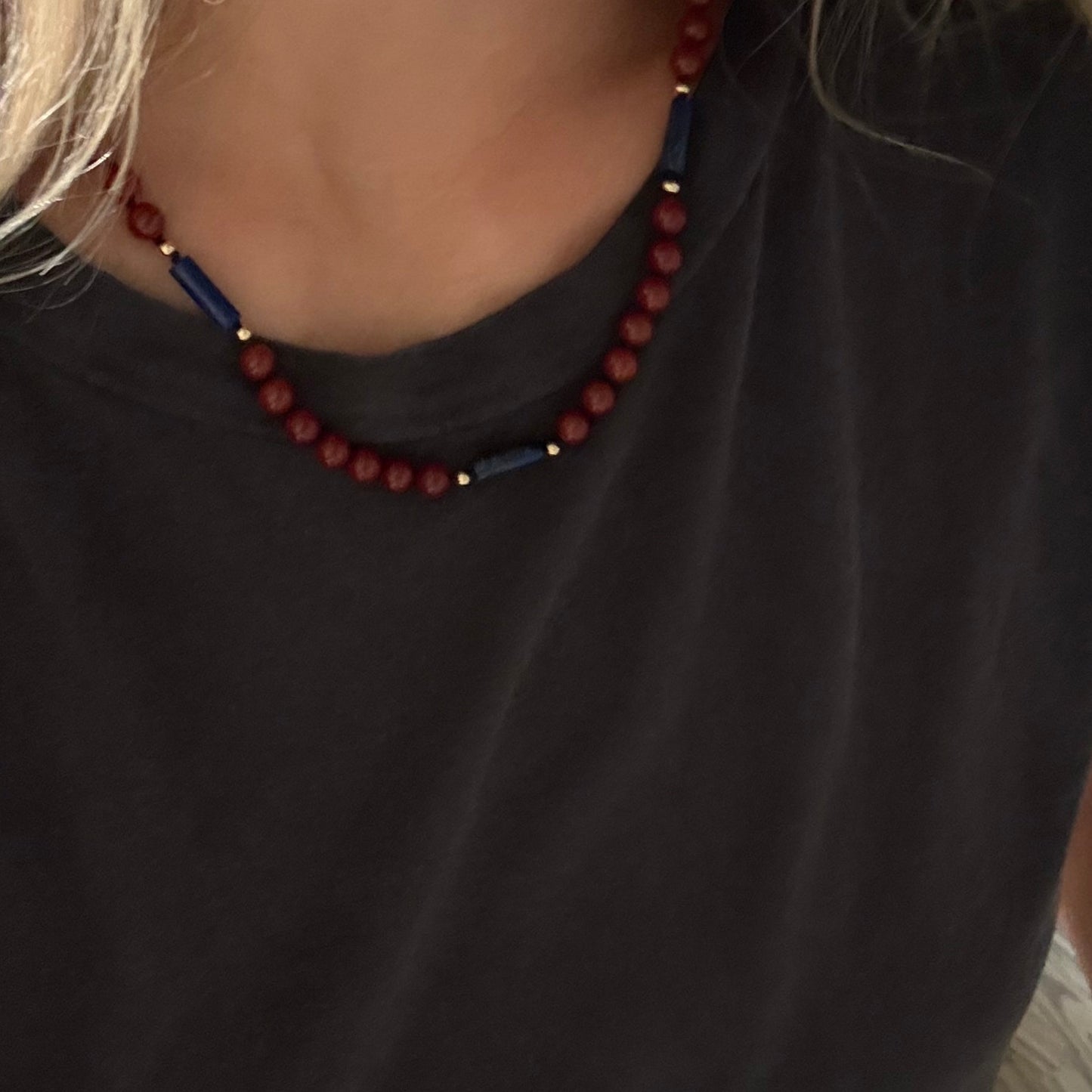 A close-up of a person wearing a beaded necklace with dark red round beads and blue cylindrical accents, paired with a dark gray t-shirt.