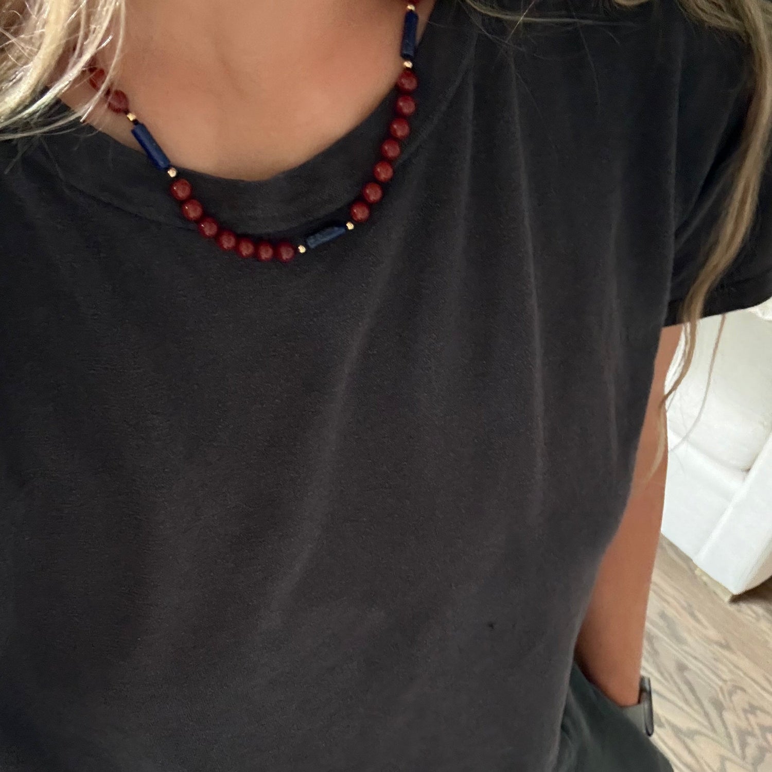 A close-up of a person wearing a dark charcoal gray t-shirt and gray drawstring pants, accessorized with a beaded necklace featuring red and navy blue beads.