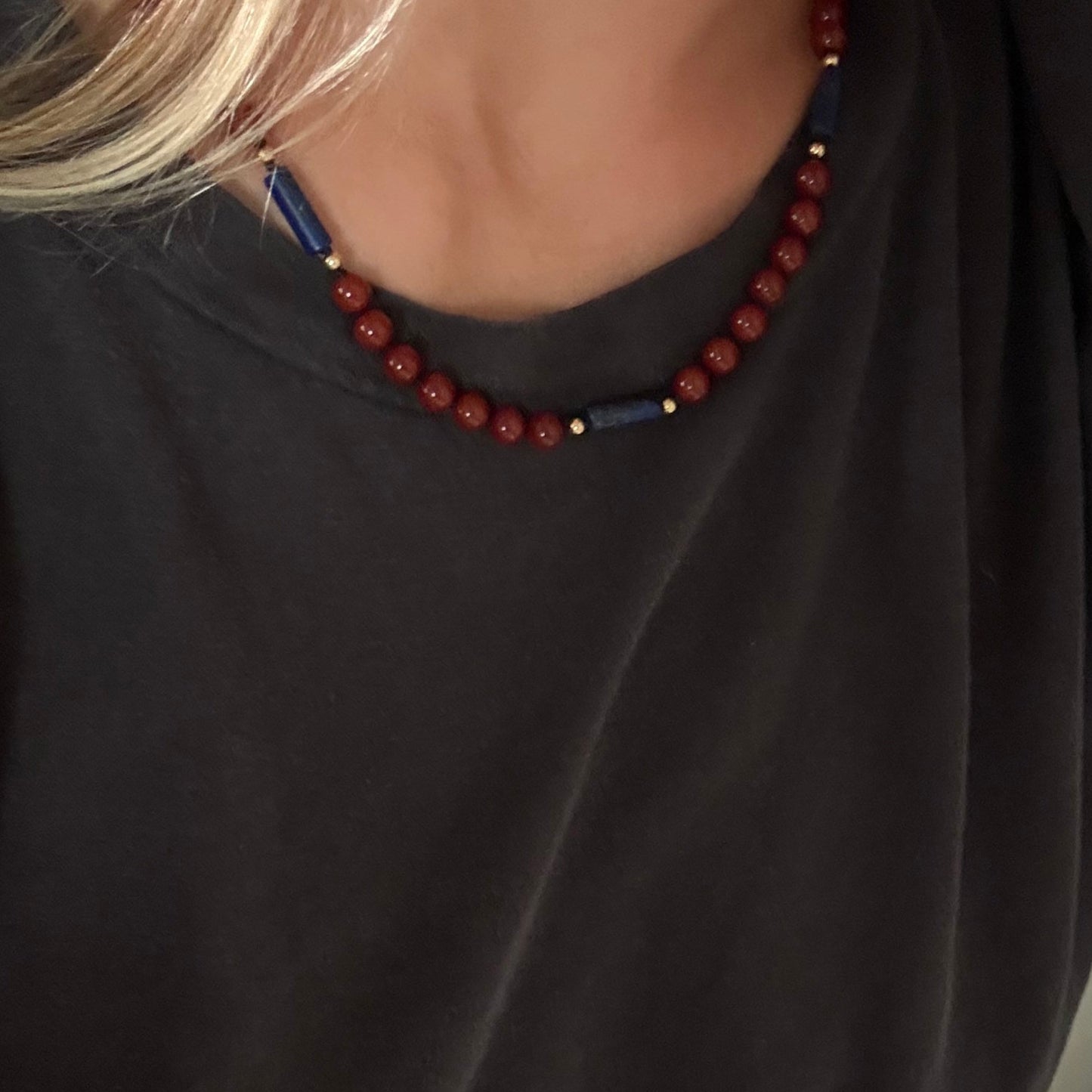 A close-up of a person wearing a dark charcoal gray t-shirt and a beaded necklace with red beads and small blue accents.