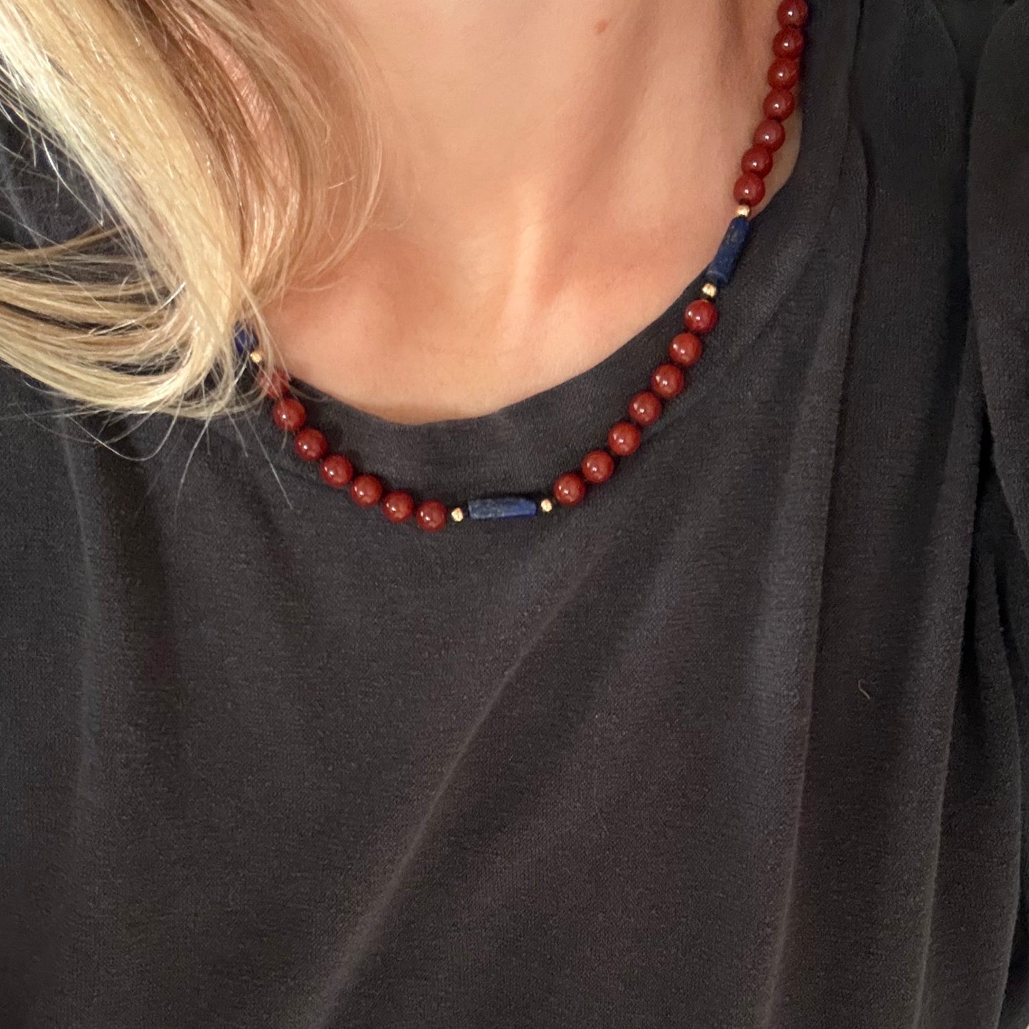 A close-up of a person wearing a beaded necklace with red round beads, a dark blue cylindrical bead, and a small gold accent, paired with a dark gray t-shirt.