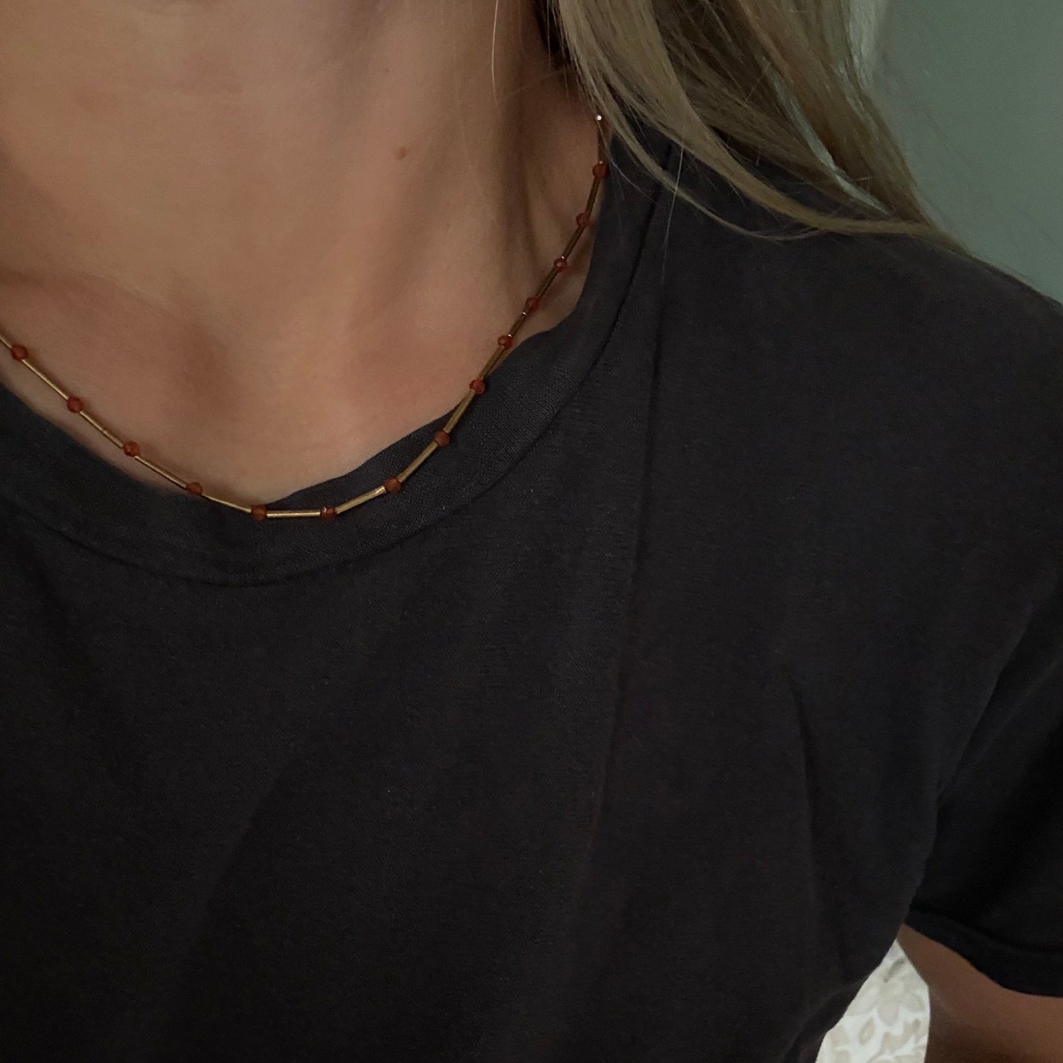 A close-up of a person wearing a thin beaded necklace with small red and gold beads, paired with a black t-shirt.