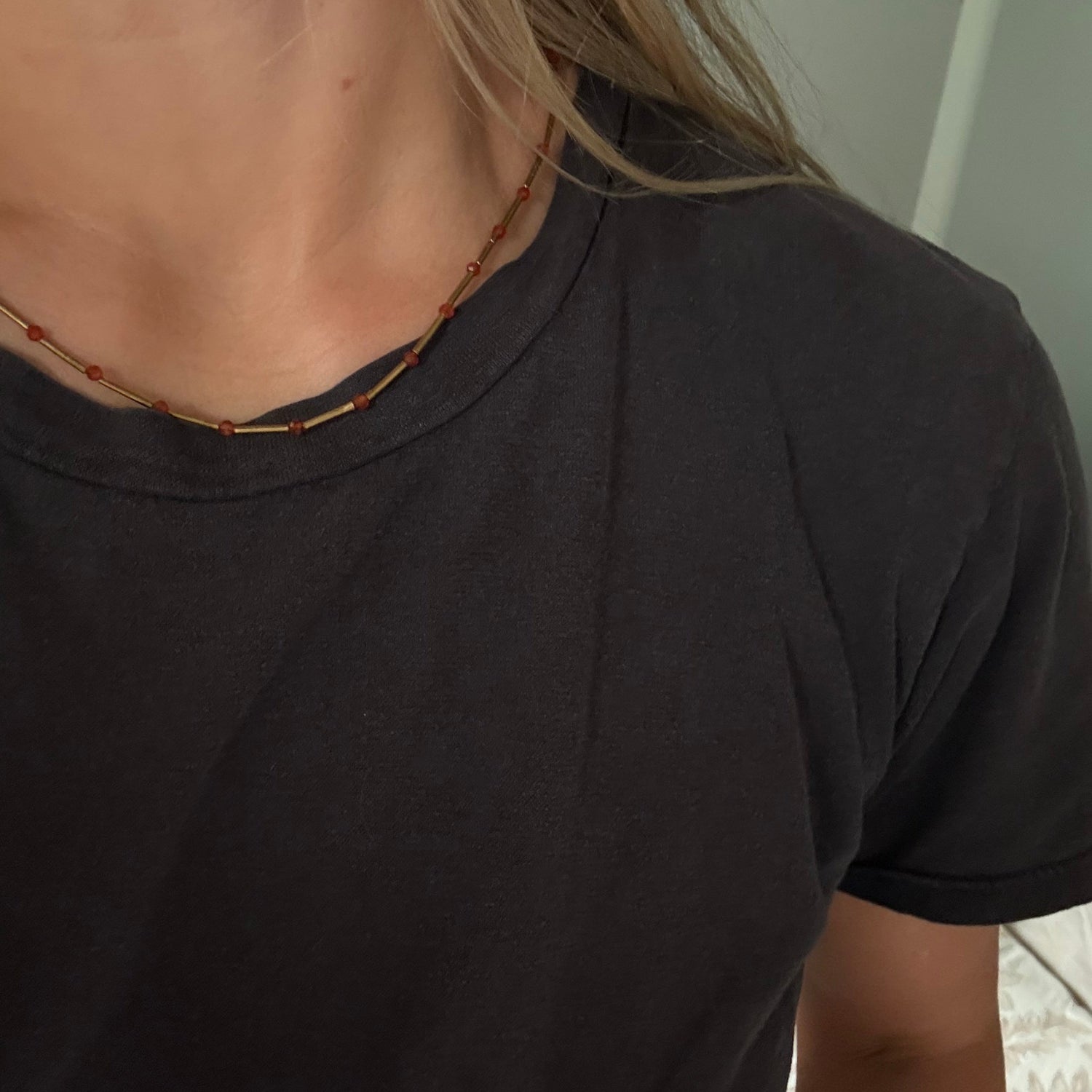 Close-up of a person wearing a black short-sleeve crewneck t-shirt and a thin beaded necklace with red and gold beads.