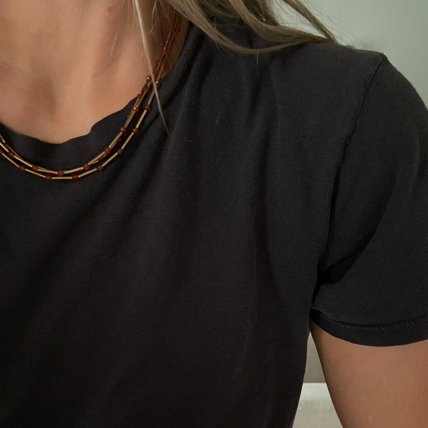 Close-up of a person wearing a black short-sleeve crewneck t-shirt and a layered beaded necklace with brown and gold beads.