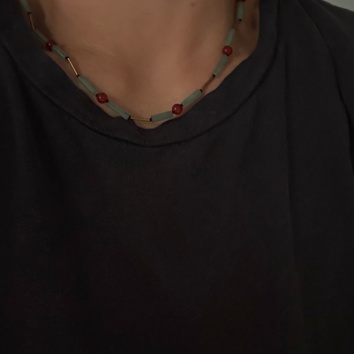 A close-up of a person wearing a black t-shirt and a beaded necklace with light blue-green beads and red accents.