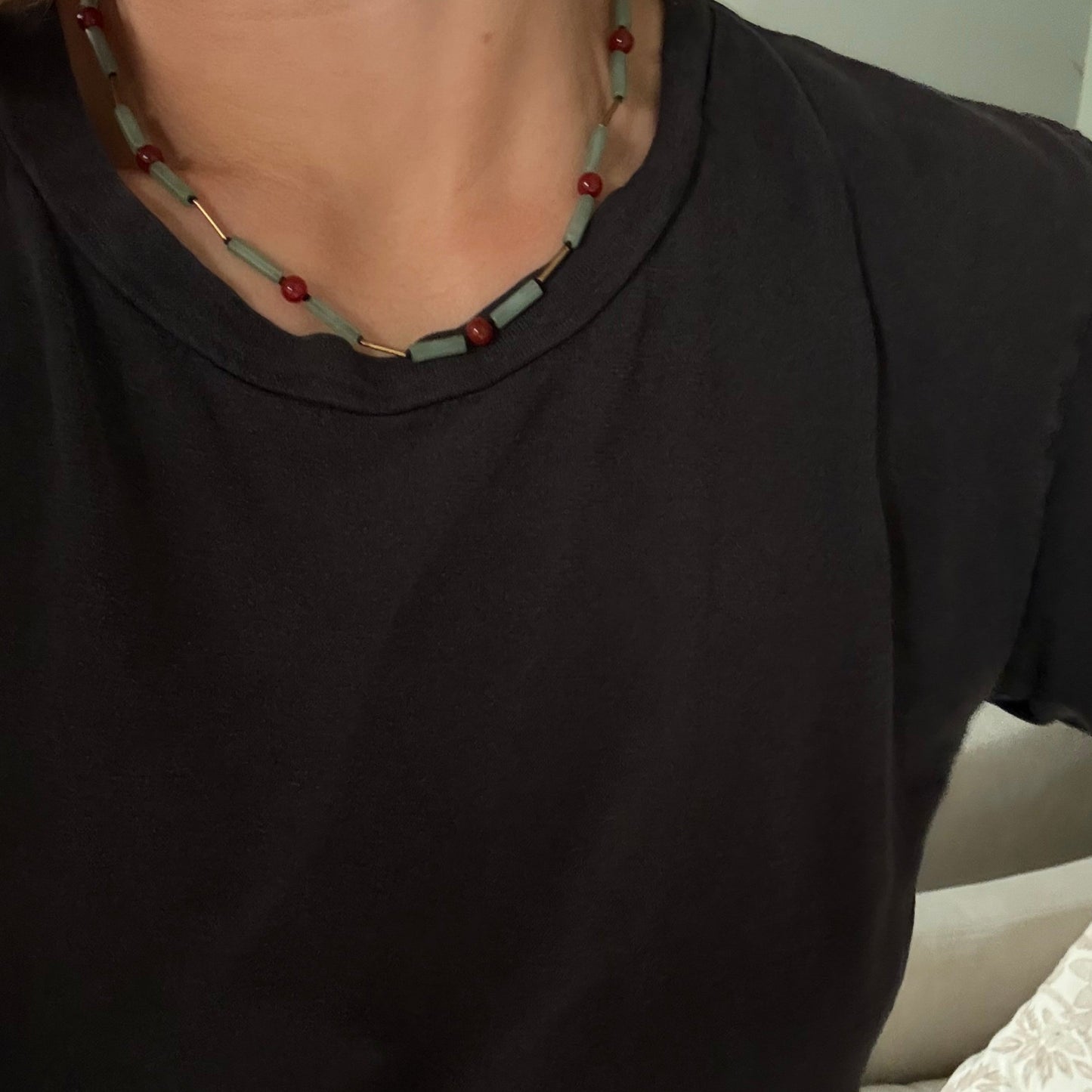 A close-up of a person wearing a black t-shirt and a beaded necklace with light blue-green rectangular beads, red round beads, and small gold-toned spacer beads.