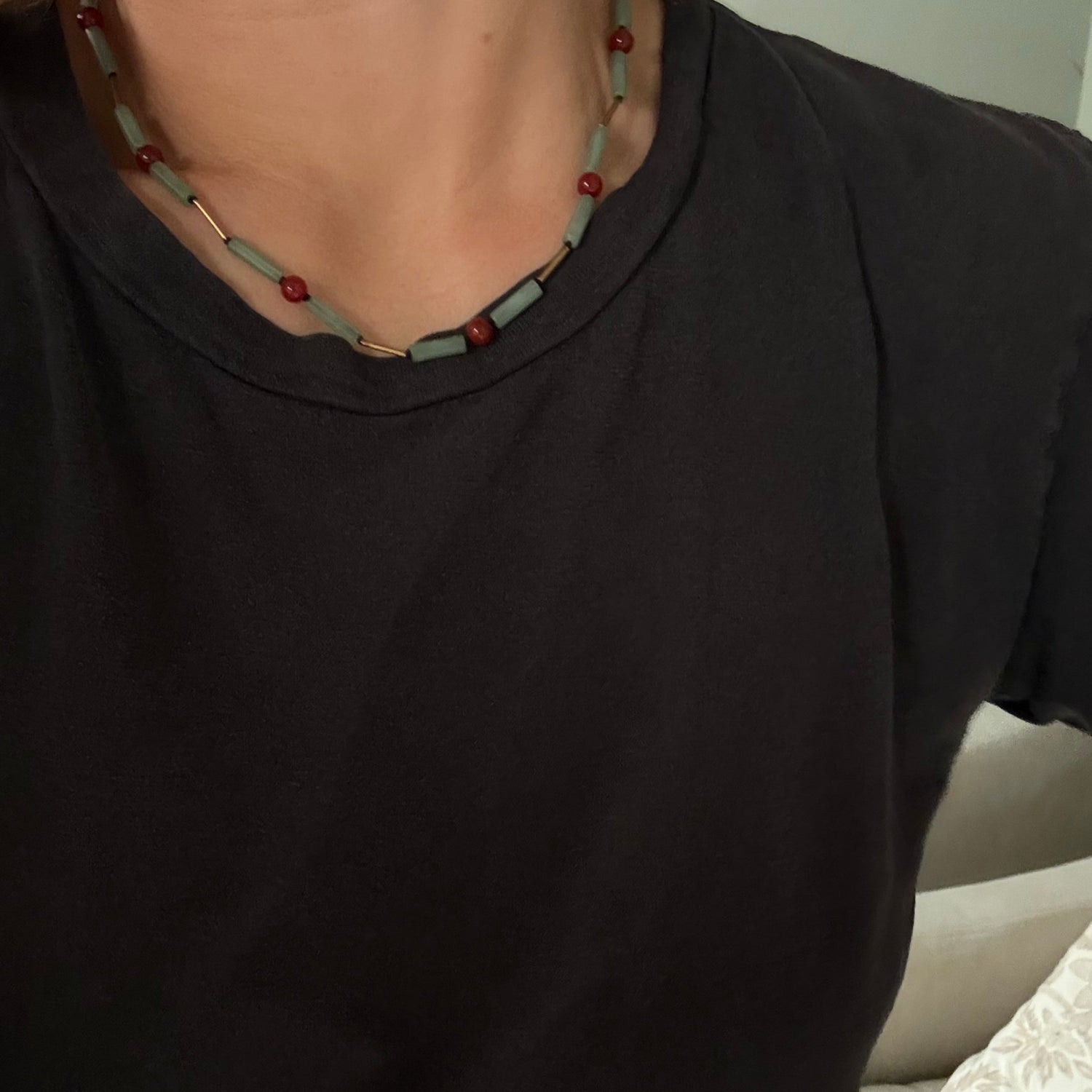 A close-up of a person wearing a black t-shirt and a beaded necklace with light blue-green rectangular beads, red round beads, and small gold-toned spacer beads.