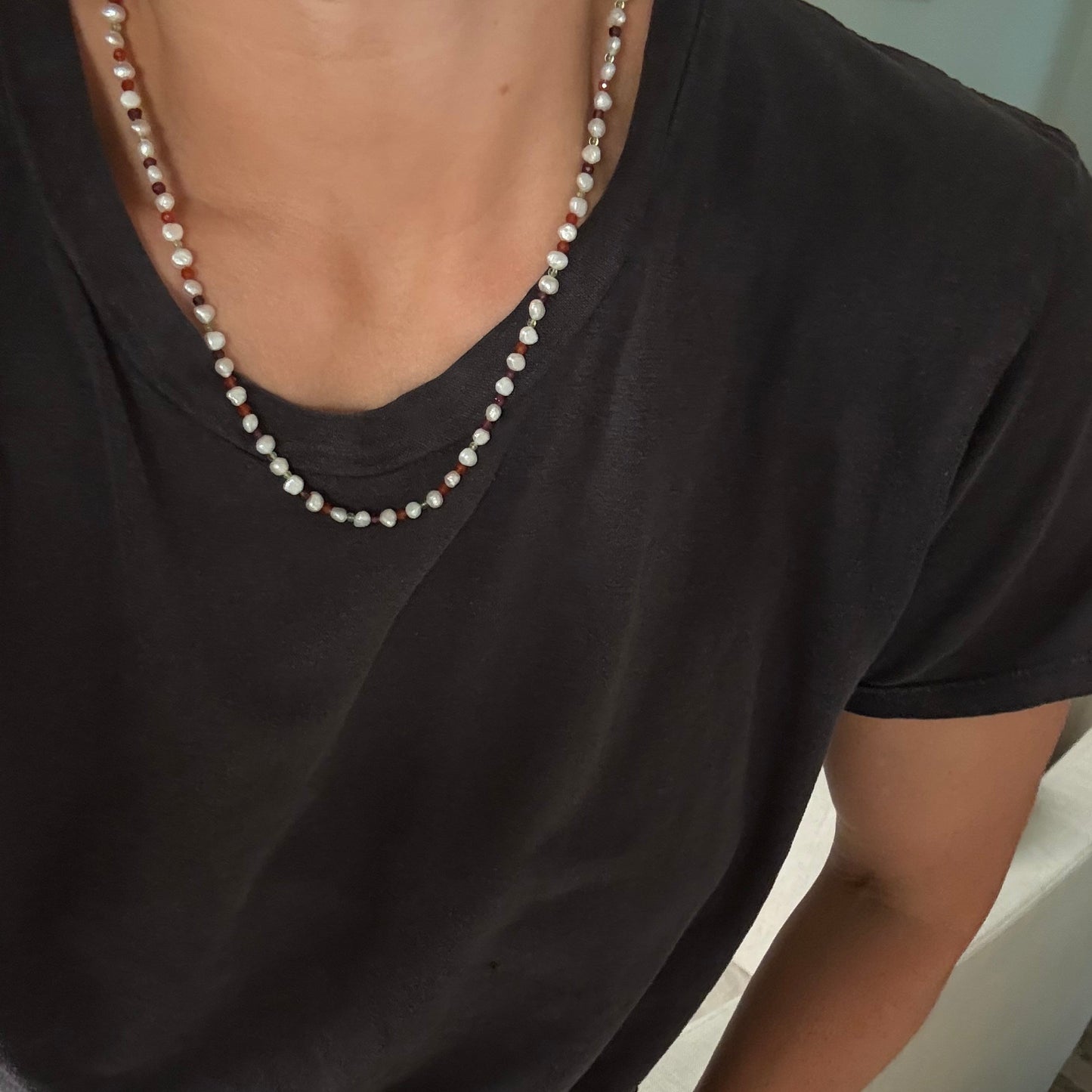 A close-up of a person wearing a short-sleeve black t-shirt and a delicate beaded necklace made of white and light pink pearls.