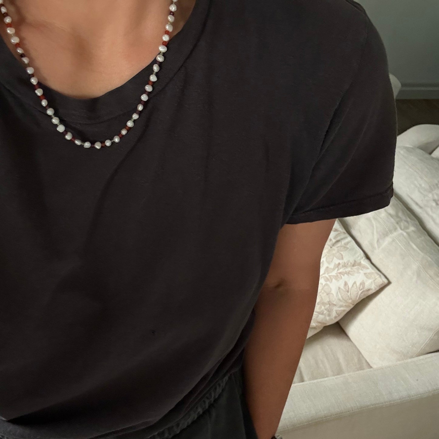 A close-up lifestyle shot of a person wearing a dark charcoal gray t-shirt and a double-strand pearl necklace with small red beads, sitting in front of a white sofa.