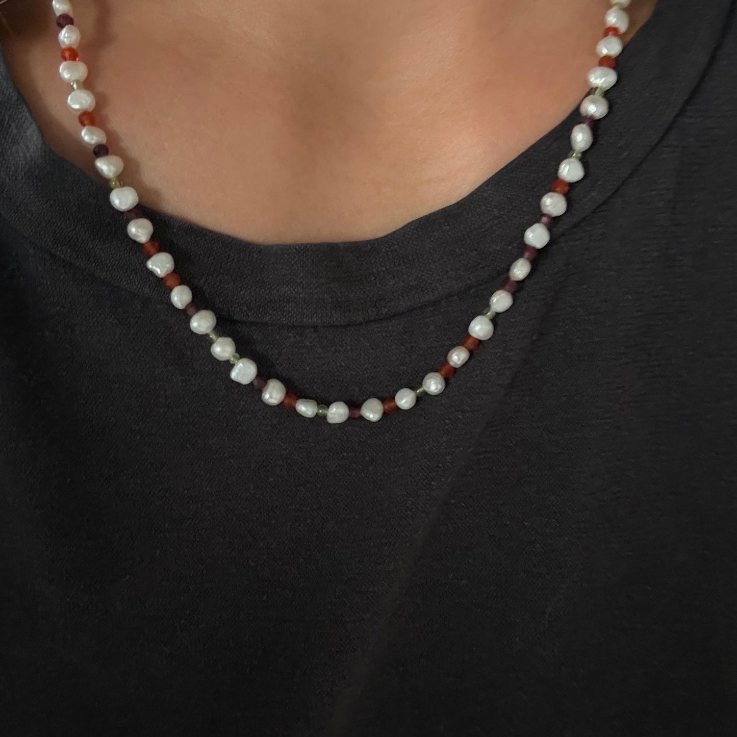 A close-up of a person wearing a double-strand beaded necklace with white baroque pearls and small reddish-brown beads, paired with a black t-shirt.