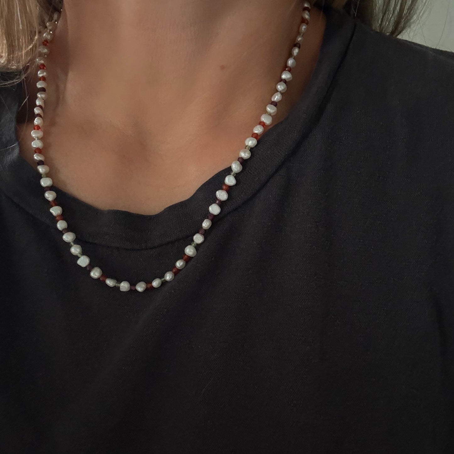 A close-up of a person wearing a beaded necklace with white and reddish-brown beads over a black shirt.