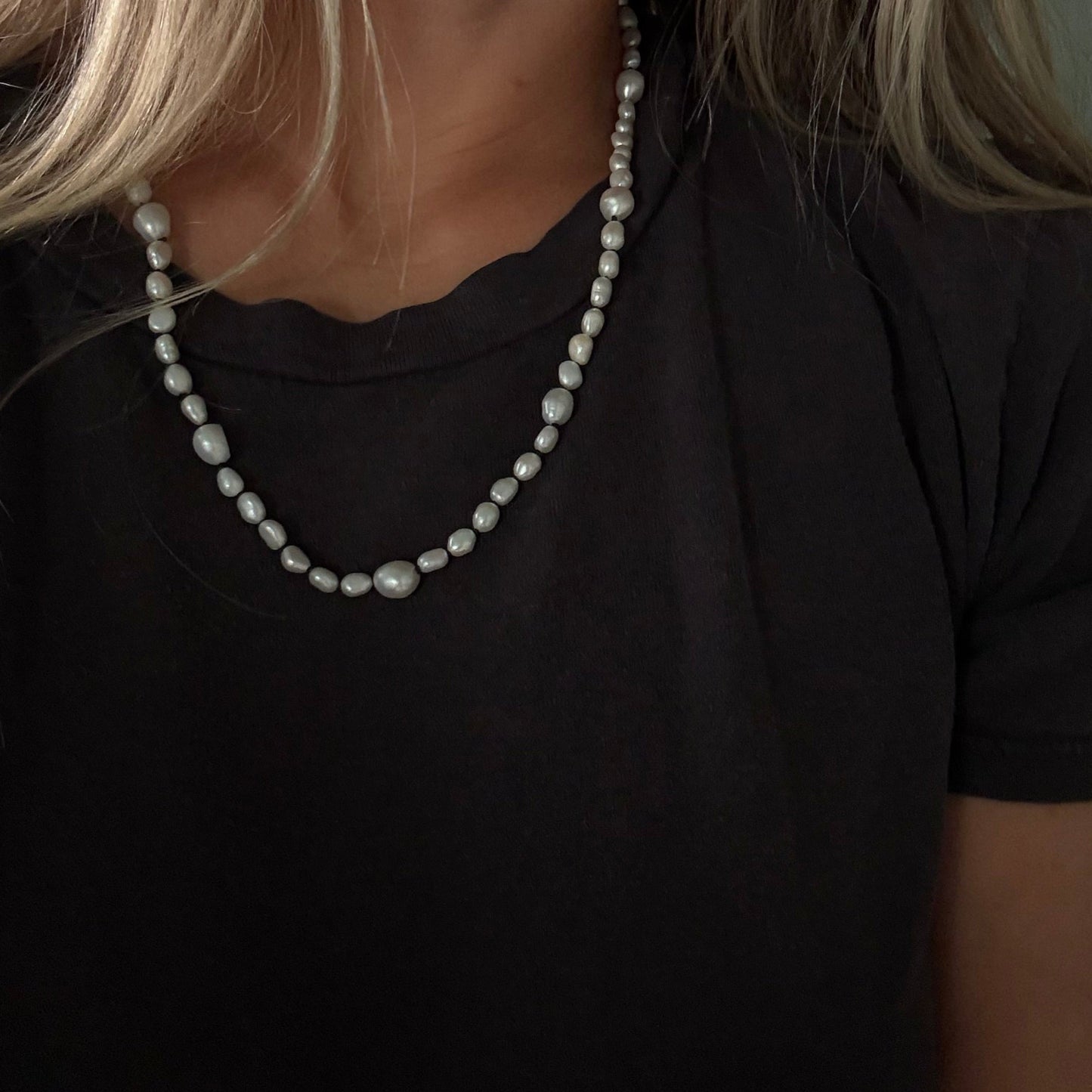 A close-up of a woman wearing a short strand pearl necklace over a black t-shirt.