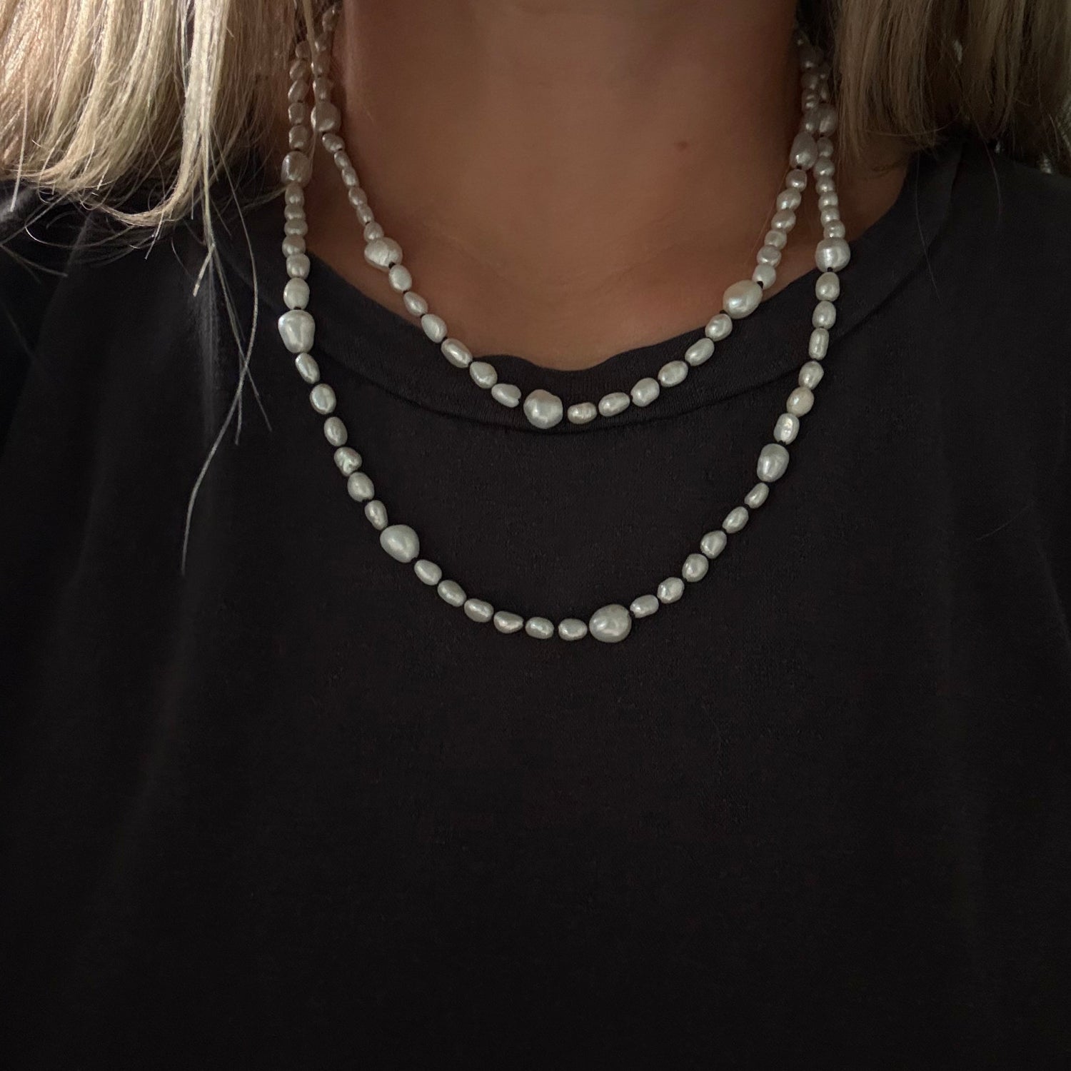 A close-up of a person wearing a double-strand necklace made of irregular white baroque pearls, styled over a black top.