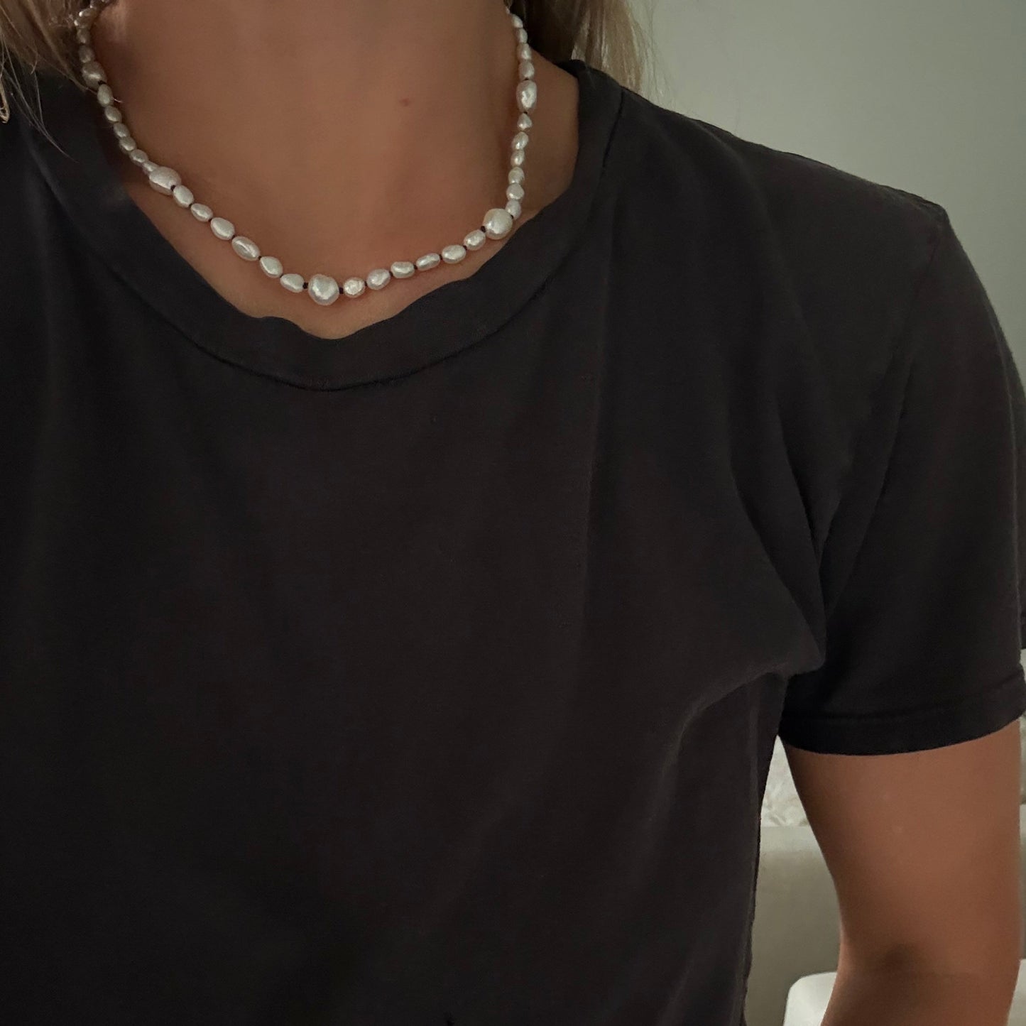 A close-up of a person wearing a black short-sleeve t-shirt and a white irregular pearl necklace.