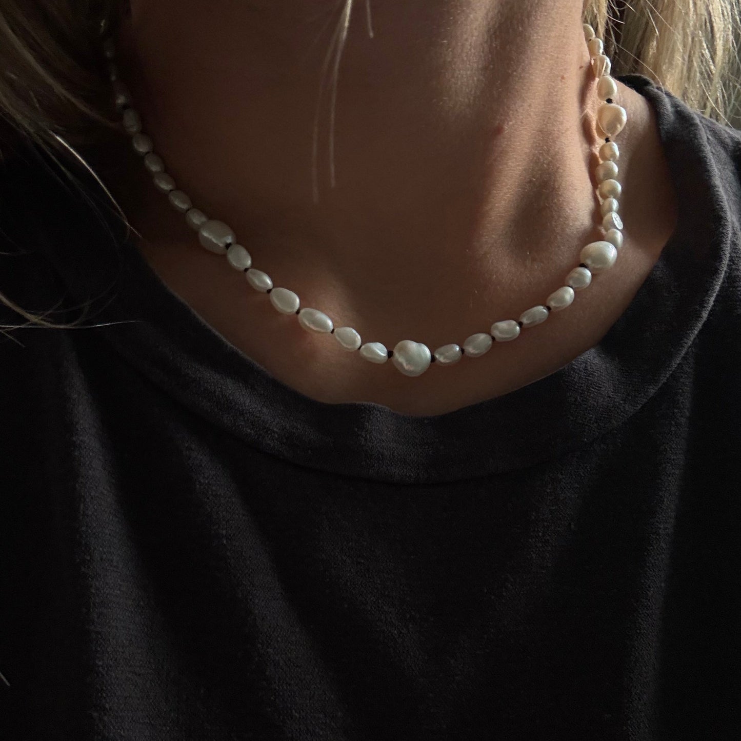 A close-up of a person wearing a beaded necklace with irregular white pearls and a gold-toned clasp, paired with a black top.