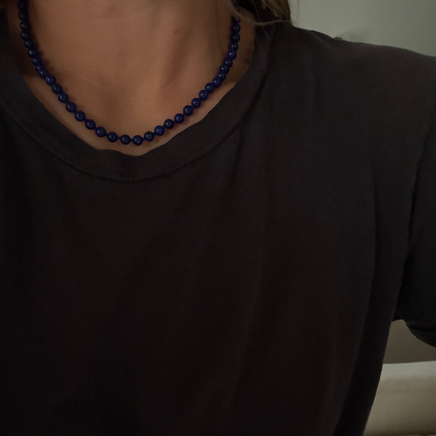 A close-up of a person wearing a dark navy blue beaded necklace with a black t-shirt in an indoor setting.