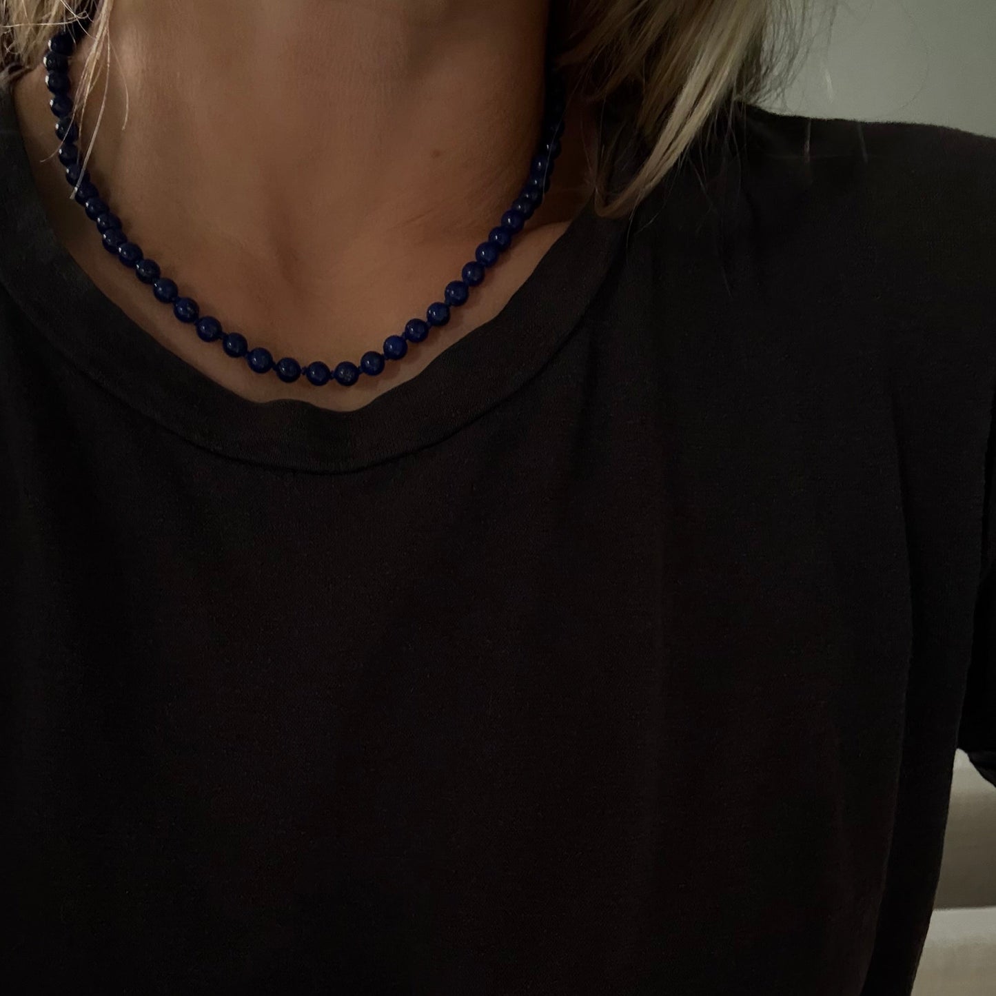 A close-up of a person wearing a dark navy blue beaded necklace with a black t-shirt, showing the necklace against the neck.
